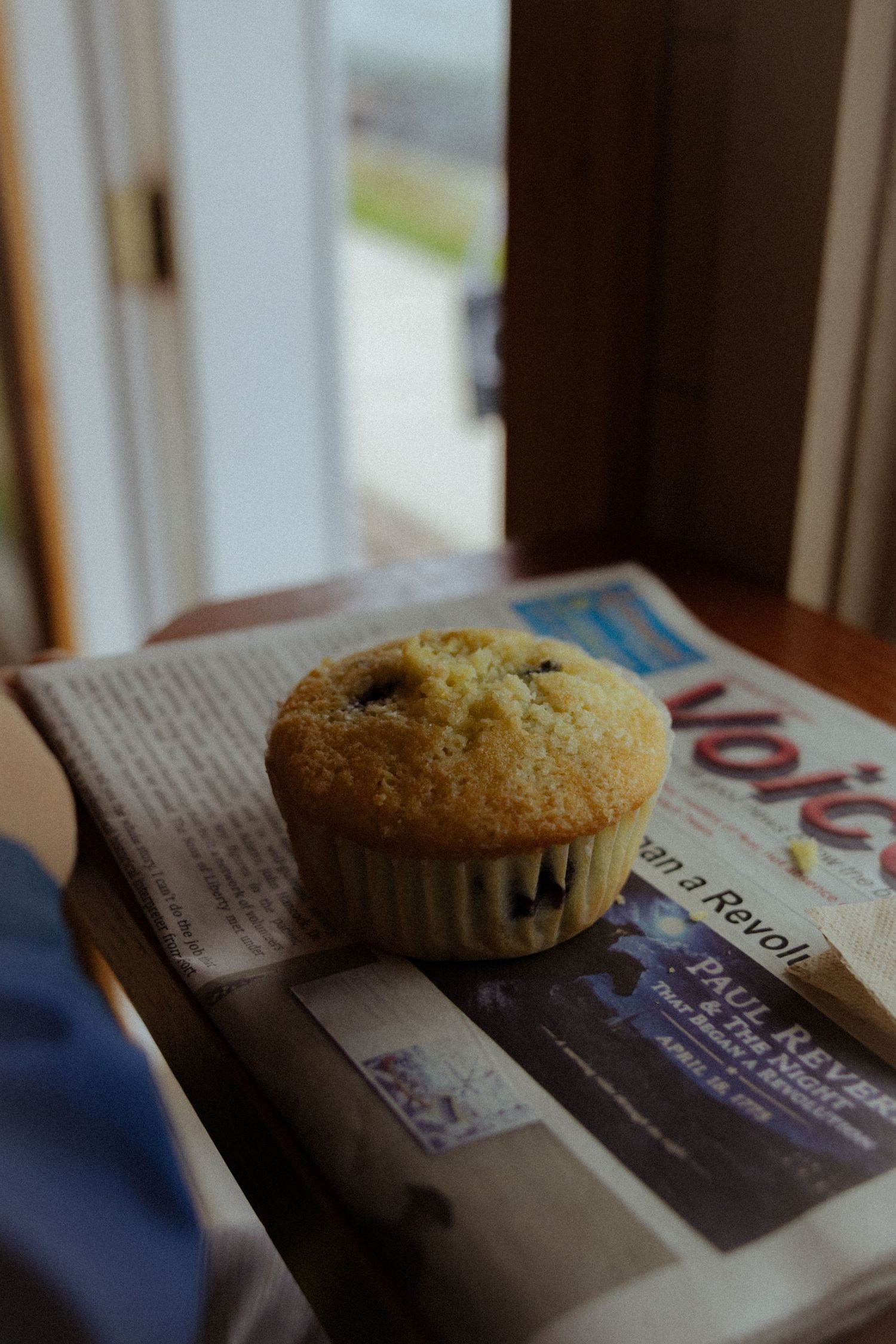 A golden blueberry muffin resting on a folded newspaper near a sunlit window.