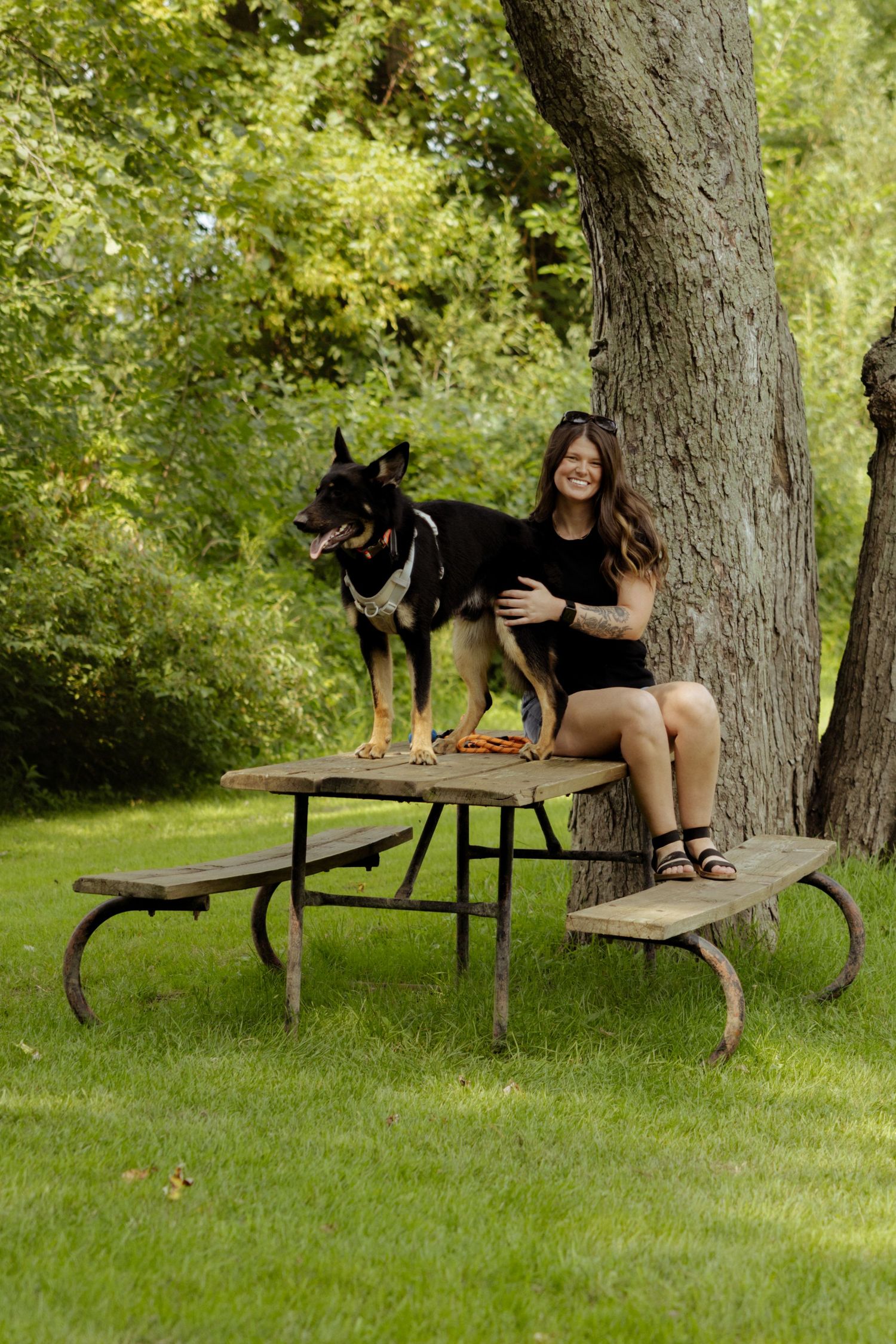 Woman sitting on a picnic table with a large black dog in a lush green park near a tree.