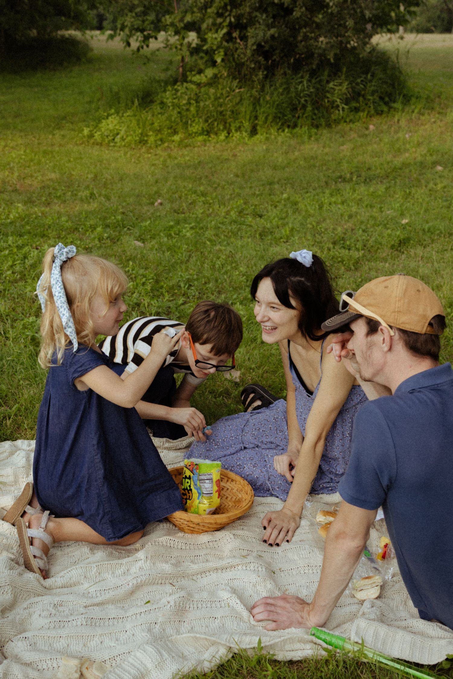 Family enjoying a sunny outdoor picnic on a blanket in a green park, with two children and parents sharing snacks together.