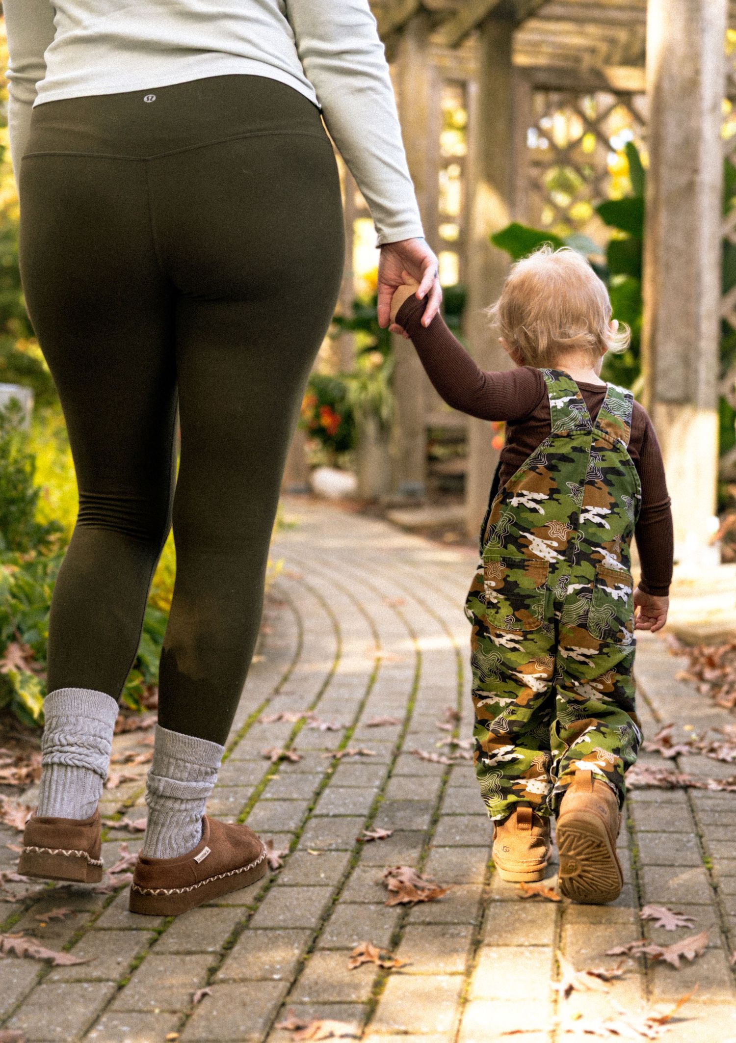 Mother and toddler walking hand in hand on a brick path, child wearing camo overalls and boots.