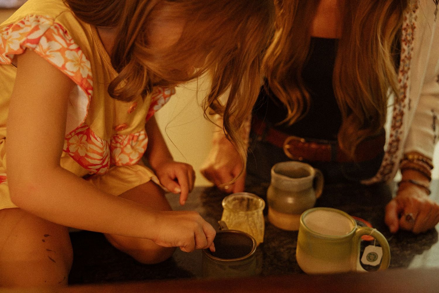 Young girl in a yellow floral top leans over a table, stirring a small cup beside handmade ceramic mugs and pots while others look on.