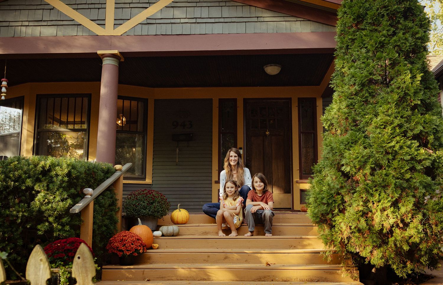 Mother and two children sit on the wooden front steps of a craftsman-style home decorated with pumpkins and autumn flowers.