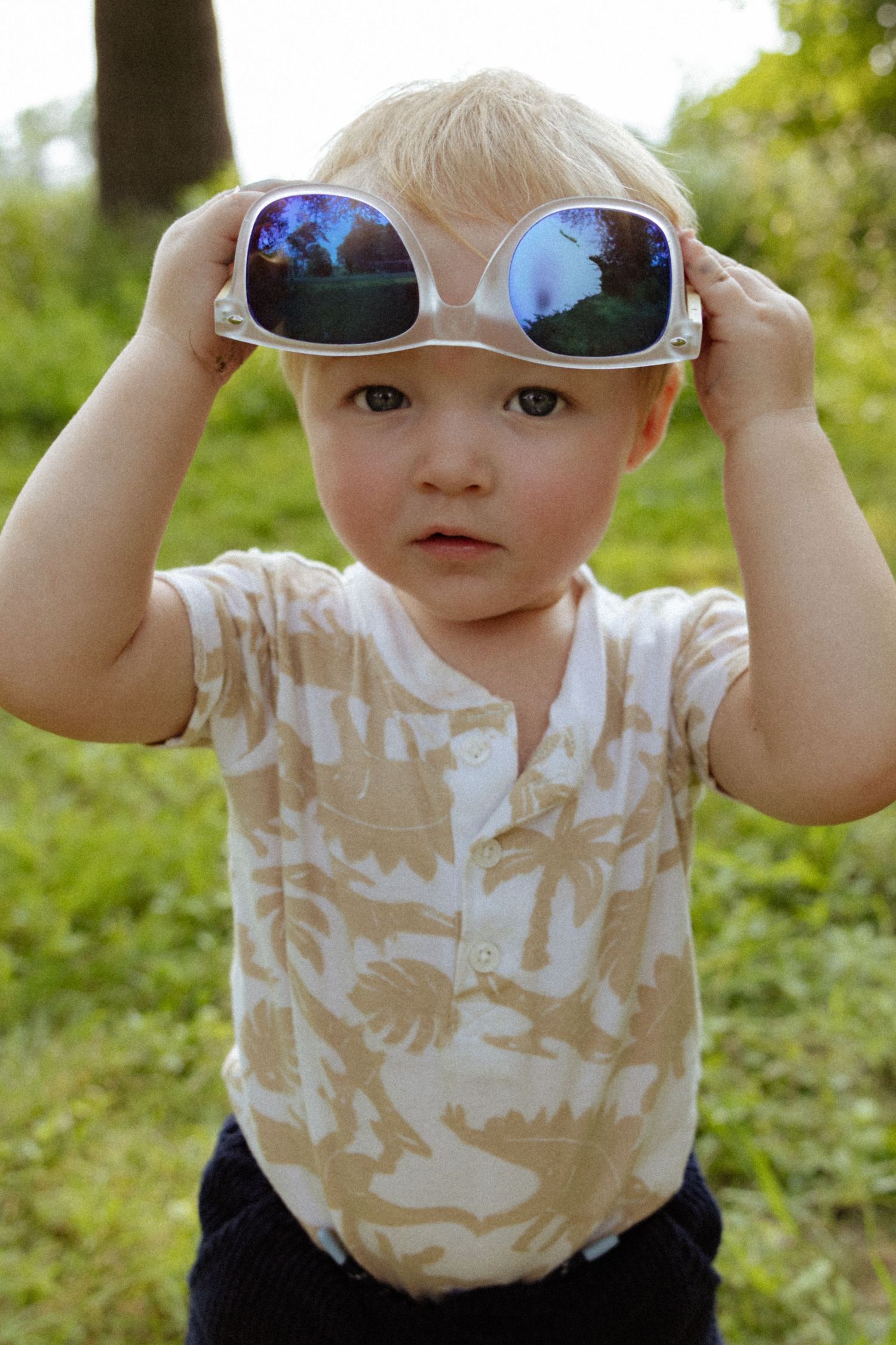 A cute blonde toddler boy wearing oversized white sunglasses outdoors in a green park setting.