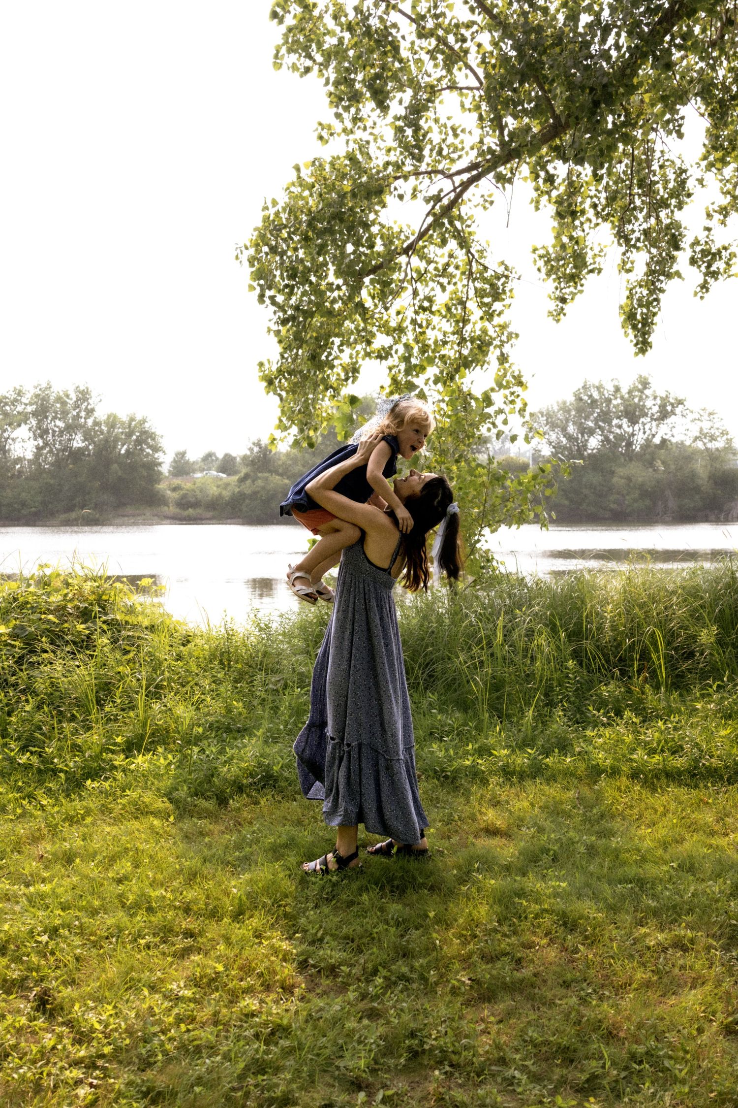 Mother lifting and hugging her child near a tree by a river in golden sunlight.