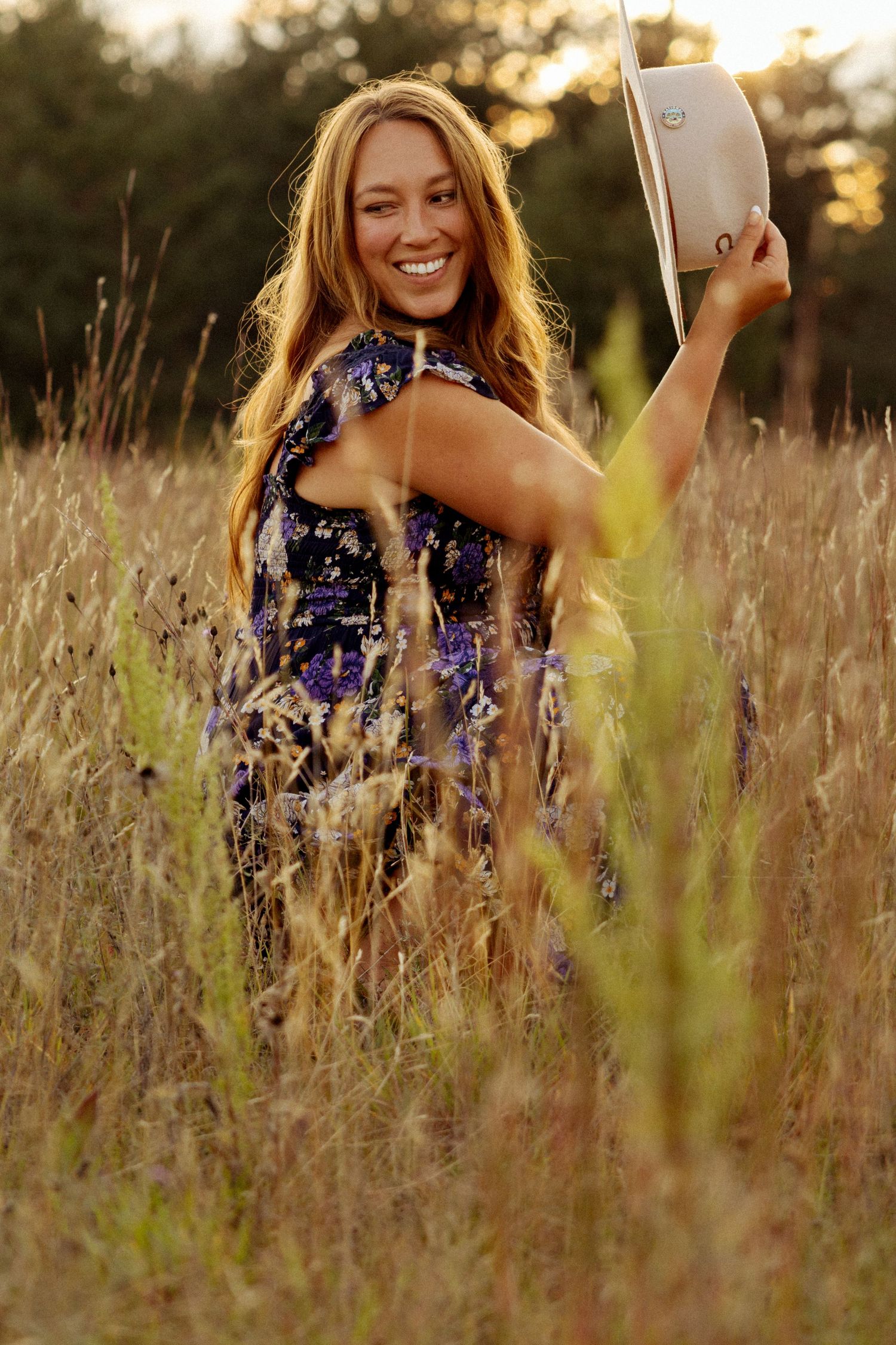 Smiling woman in floral dress holding white hat in golden field at sunset.