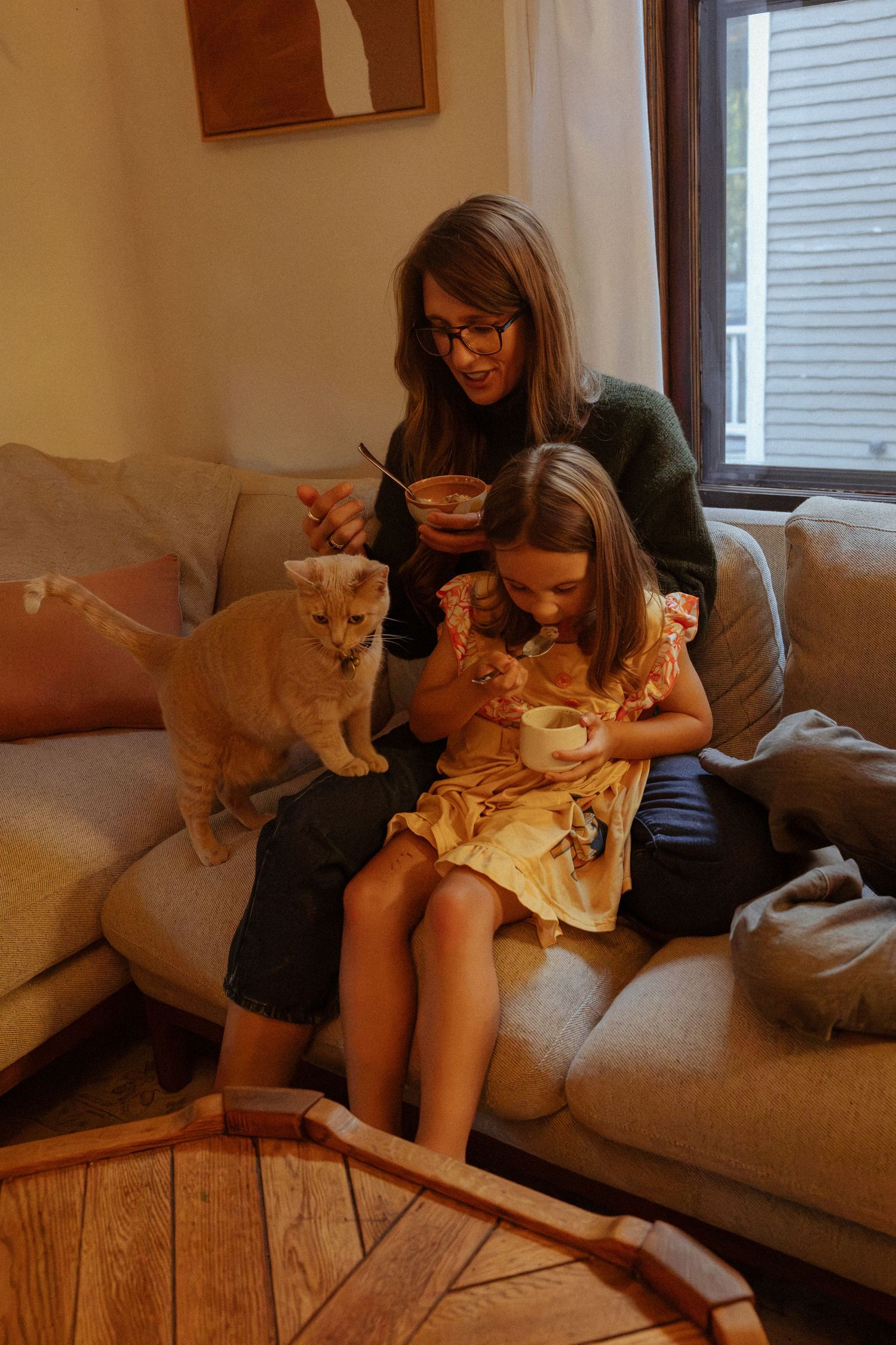 A woman with glasses and a girl in a yellow dress sit on a sofa with small bowls while an orange cat stands beside them.
