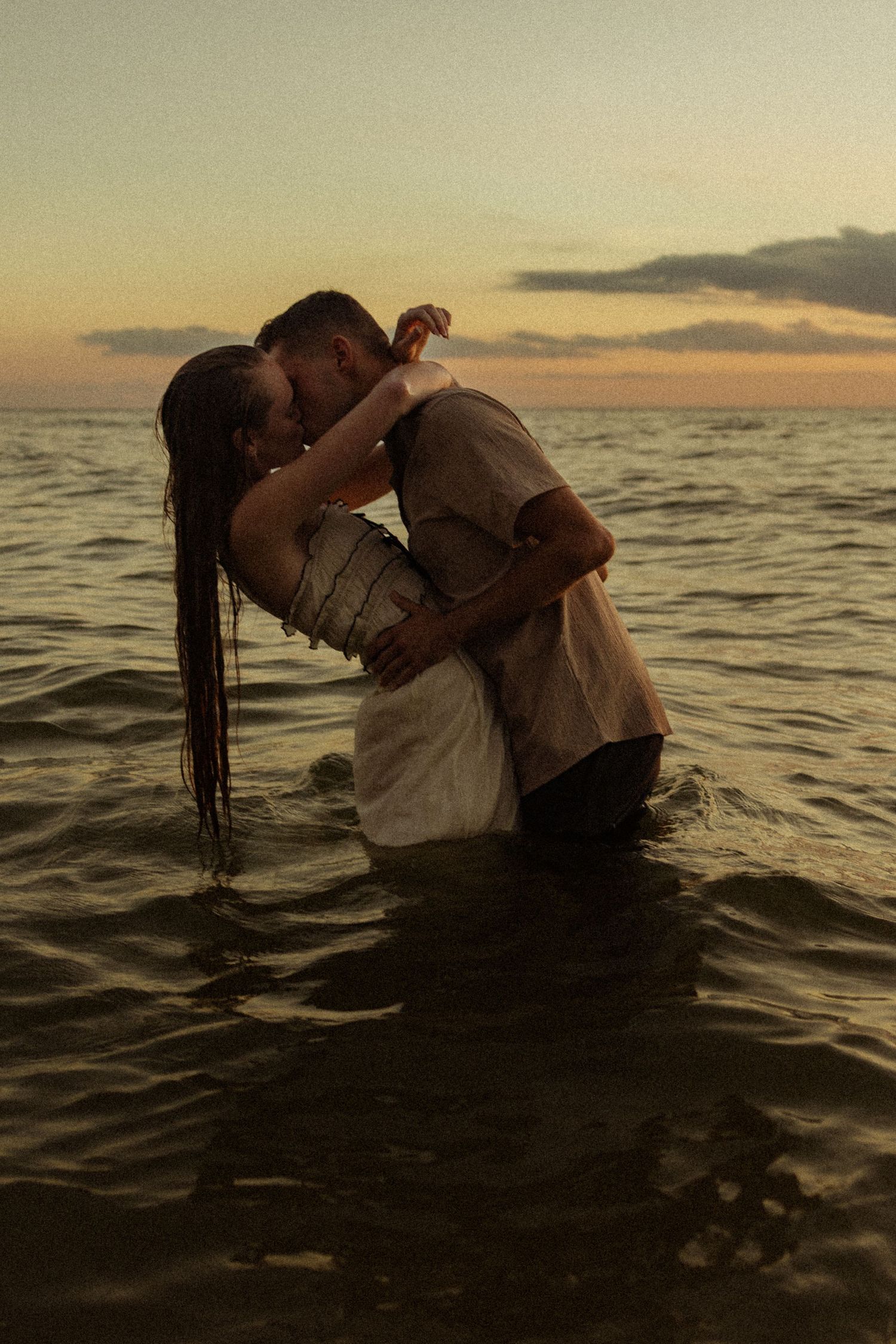Couple kissing romantically in ocean waves at sunset, wearing wet clothes, embracing in shallow water with colorful sky.