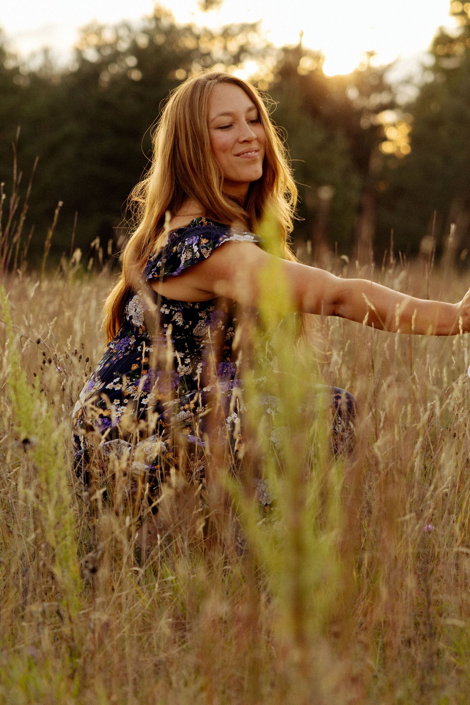 Woman in floral dress sitting in golden field at sunset, smiling with eyes closed, long hair blowing gently.