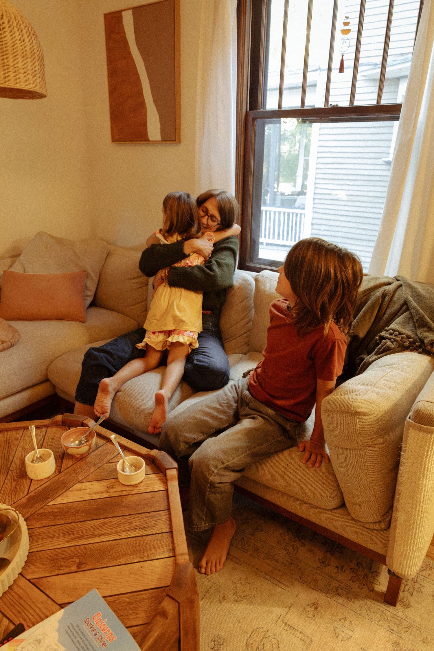 A woman hugs a young girl on a cozy sofa while a boy in a red shirt sits nearby, with a wooden coffee table in front.