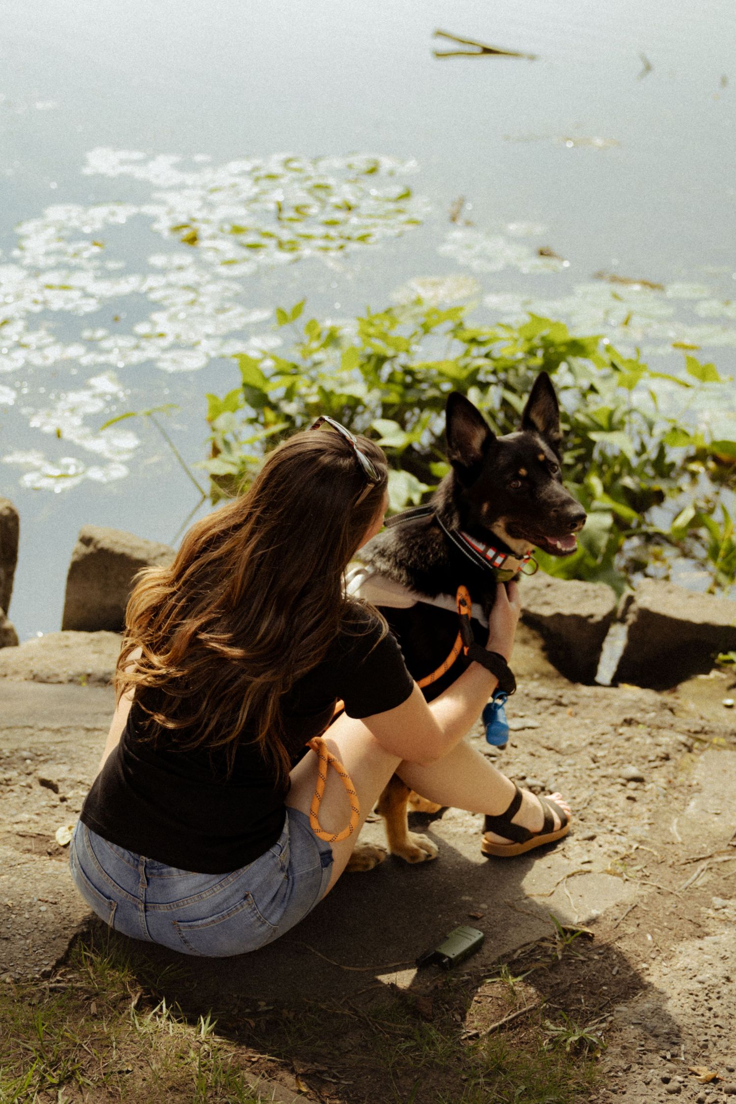 Young woman with long brown hair crouching by a pond, hugging her black dog on rocky shore.
