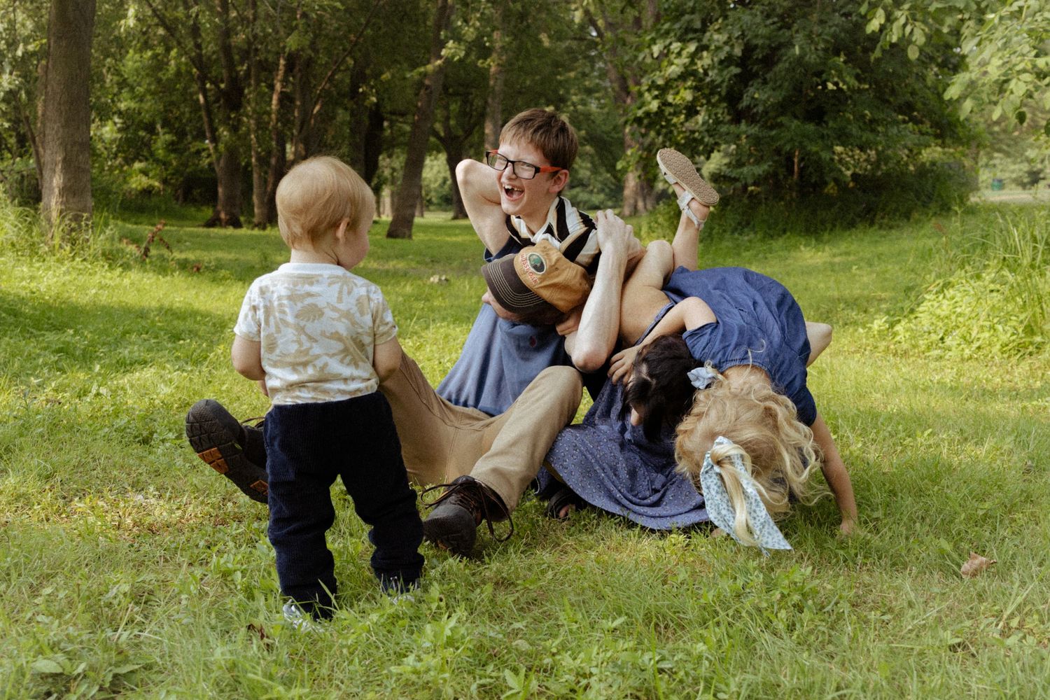 A woman playfully wrestles with a dog on green grass while a toddler watches nearby in a sunny park.