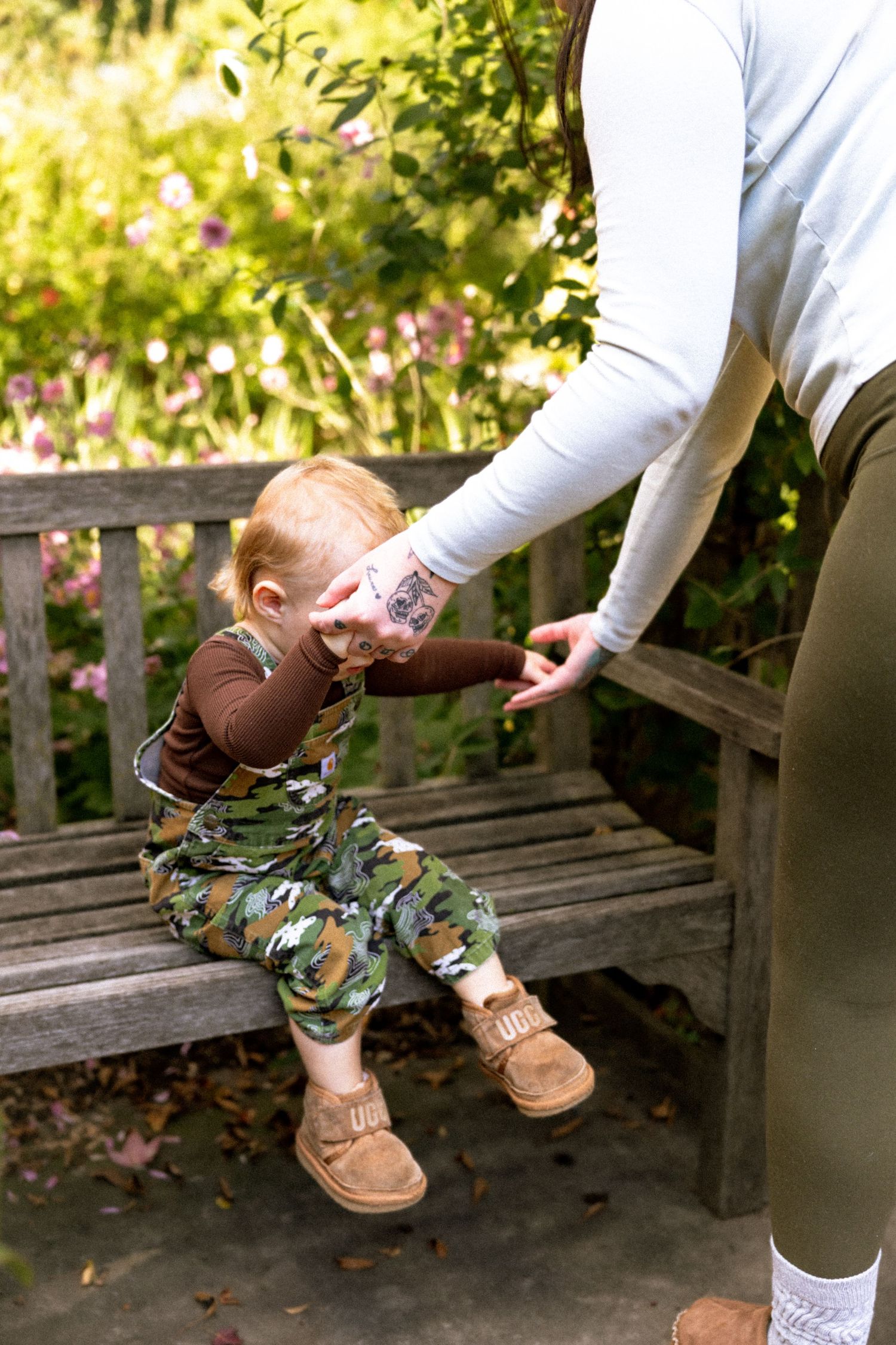 A toddler in camo overalls sits on a wooden bench, holding an adult's tattooed hand in a sunny garden.