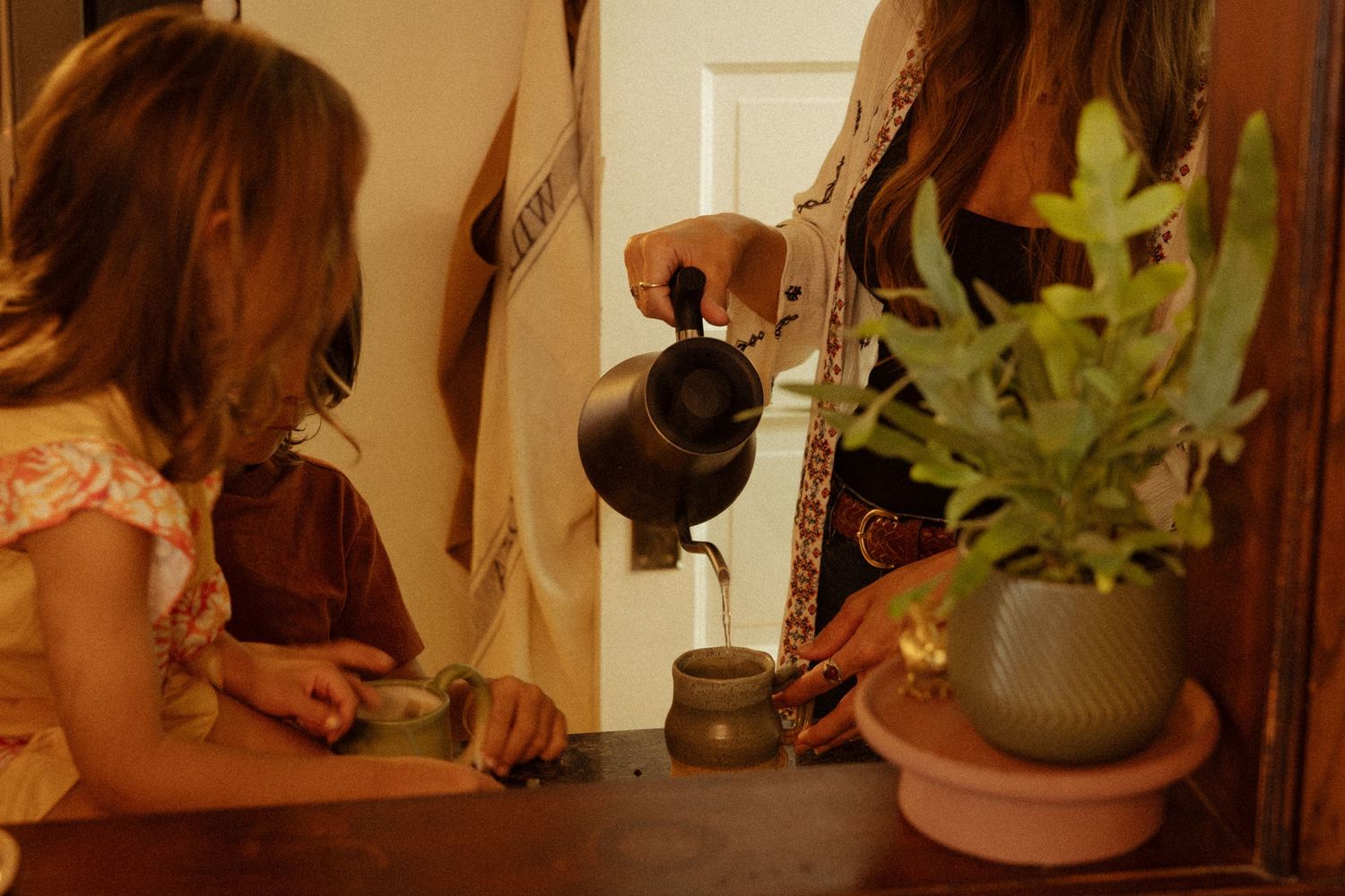 A woman pours tea from a dark kettle into a small cup while a young girl watches beside a potted green plant on a wooden table.