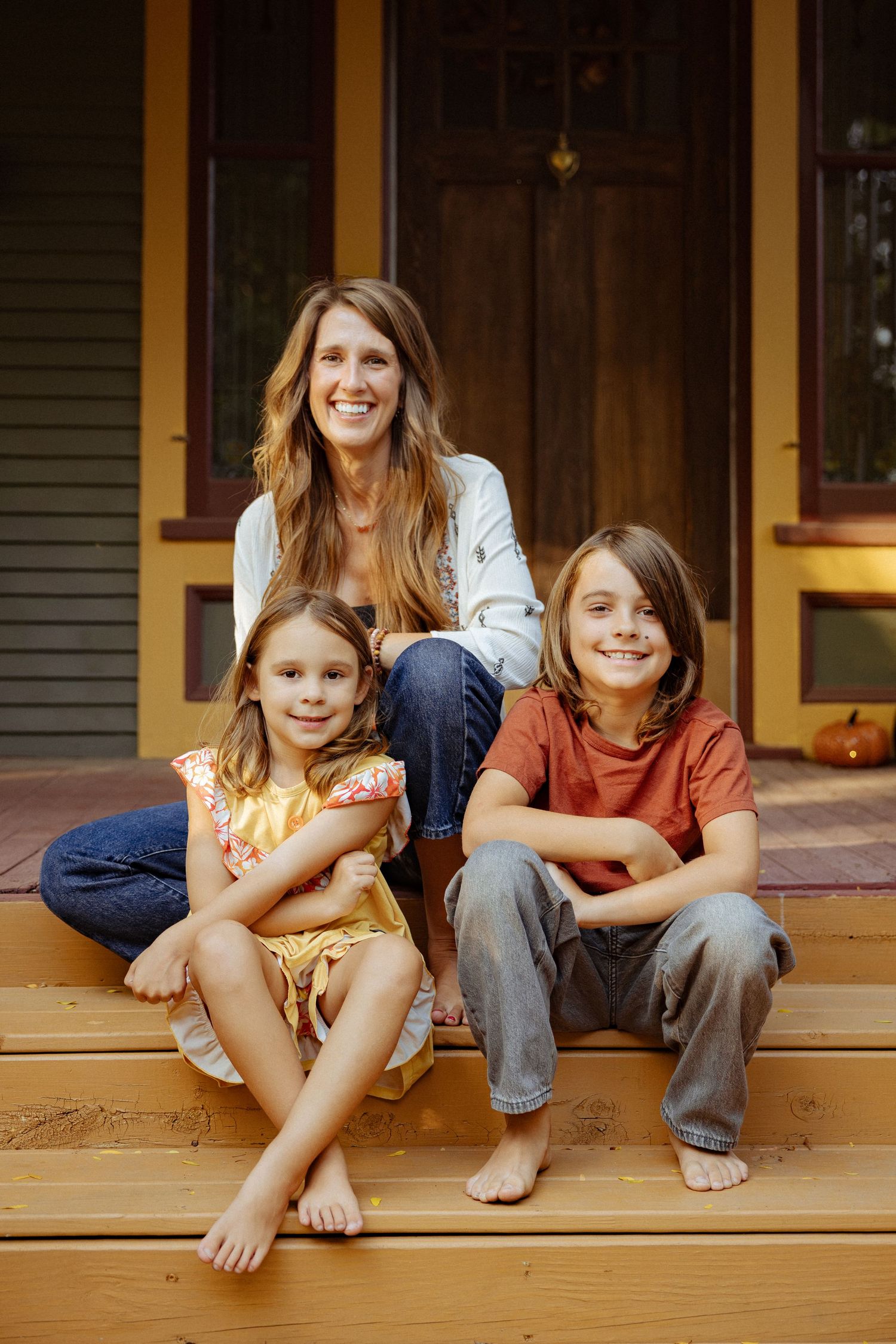 A smiling woman sits with two barefoot children on wooden porch steps in front of a rustic brown door with a small pumpkin nearby.