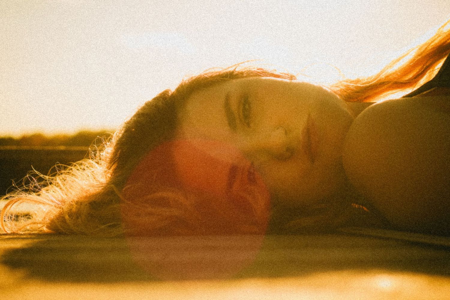 Young woman with long red hair lying down in golden sunset light, eyes closed, dreamy film photography aesthetic.