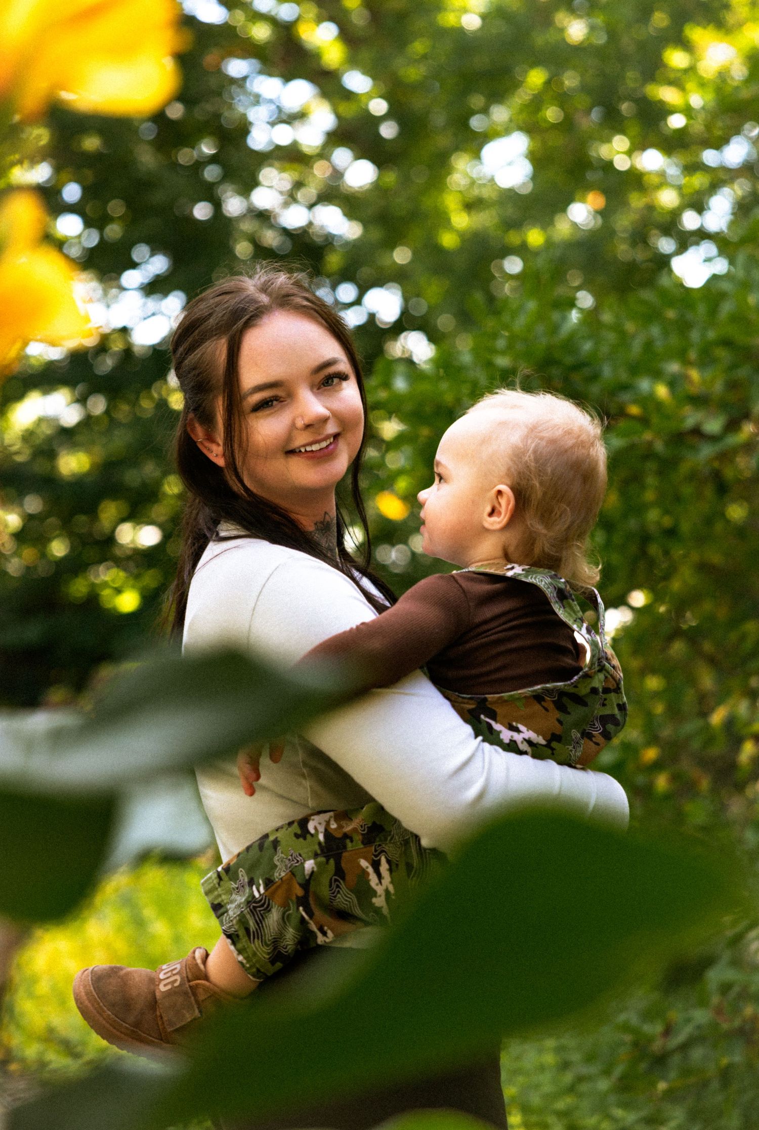 Smiling young woman holding a baby outdoors among green trees and sunflowers in a garden setting.