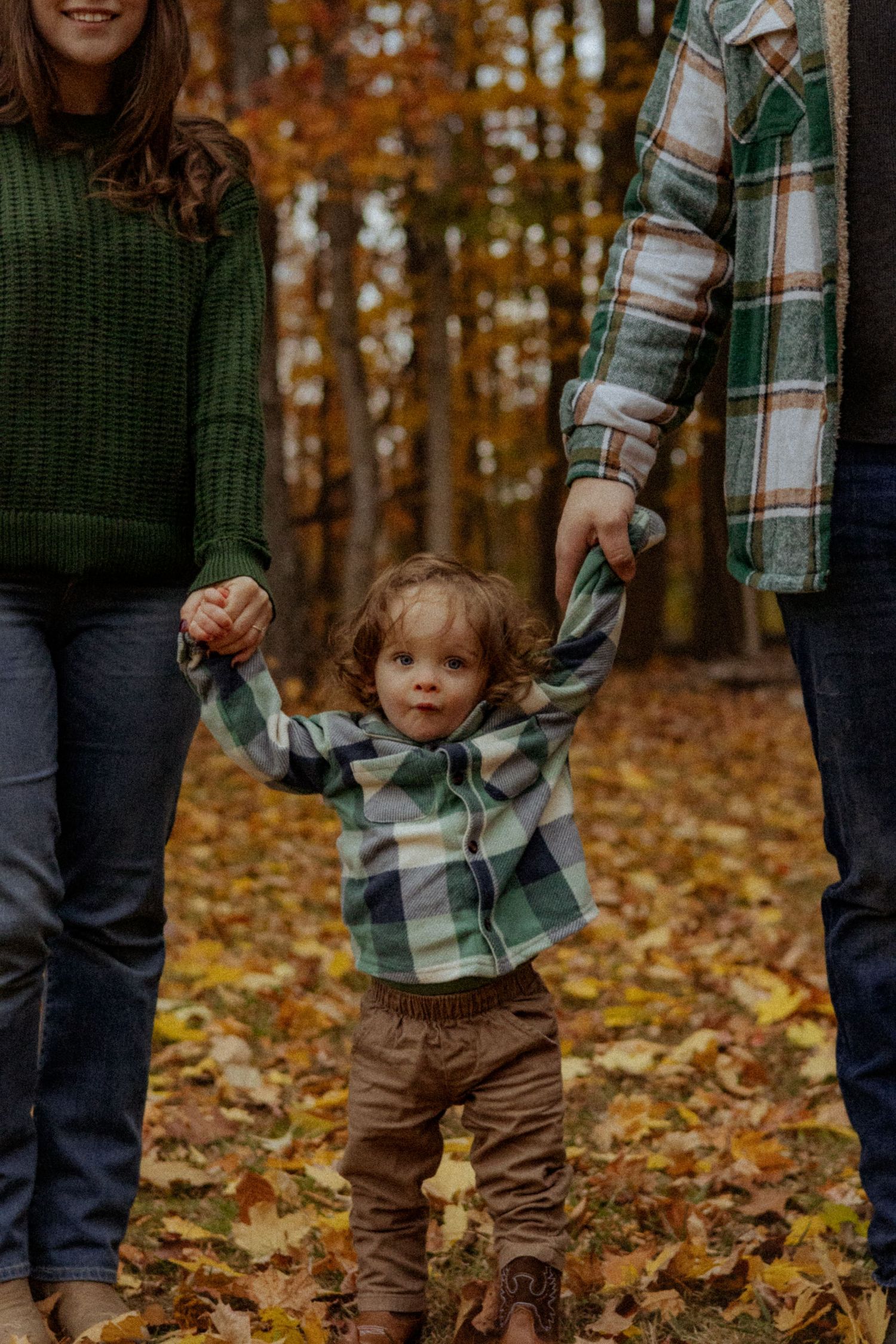 Toddler in plaid shirt holds parents hands walking through autumn forest with colorful fallen leaves.