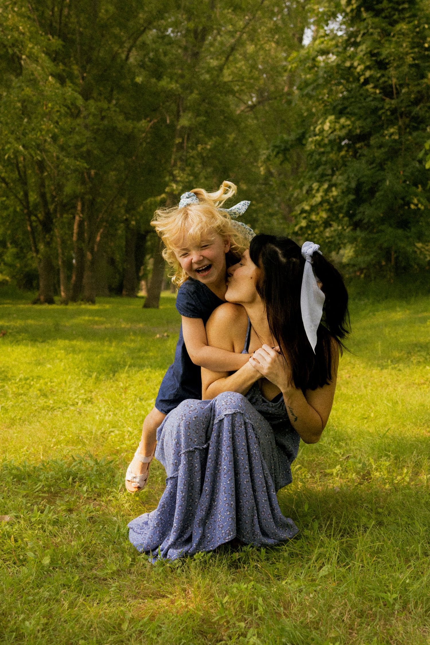 A mother crouches on green grass hugging her laughing blonde toddler daughter in a sunny park.