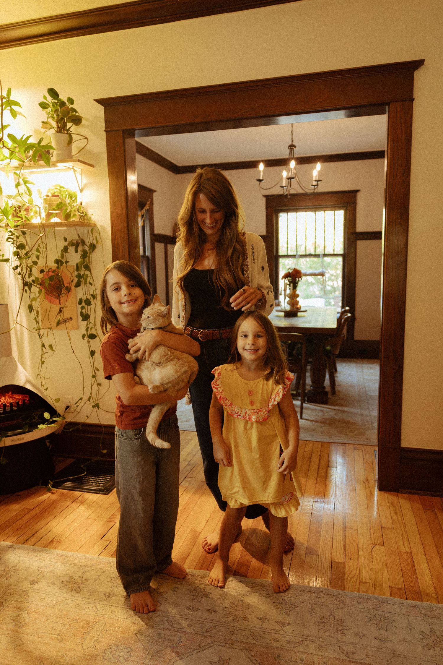 A smiling woman stands behind two barefoot children, one holding a cat, in a bright hallway with wood floors, trim and a dining room beyond.
