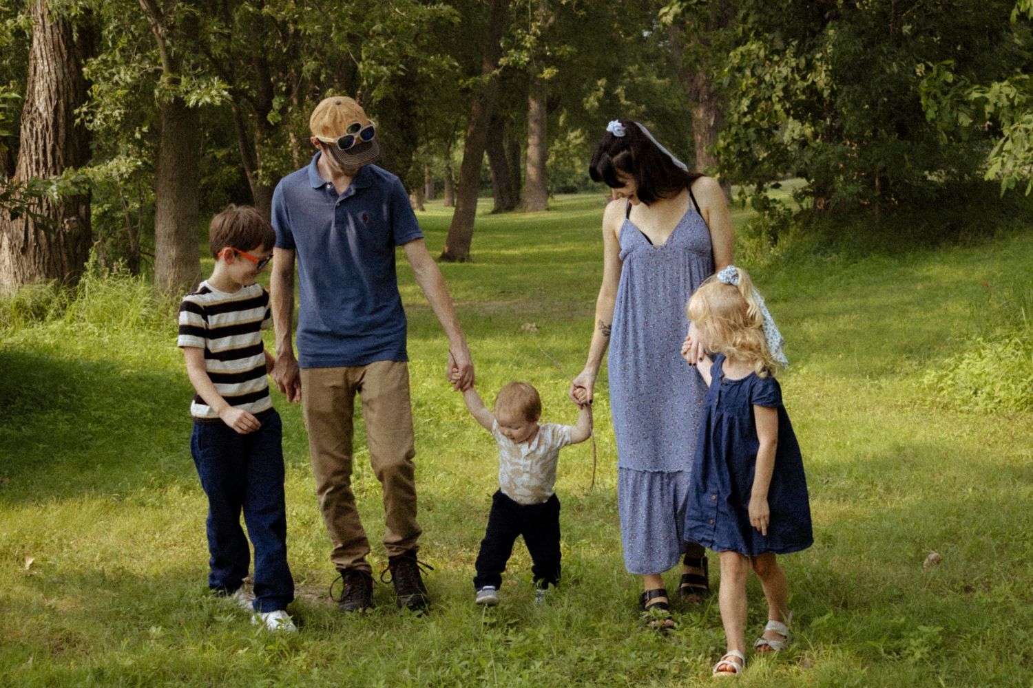 A family of five walking together through a lush green tree-lined park on a sunny day.