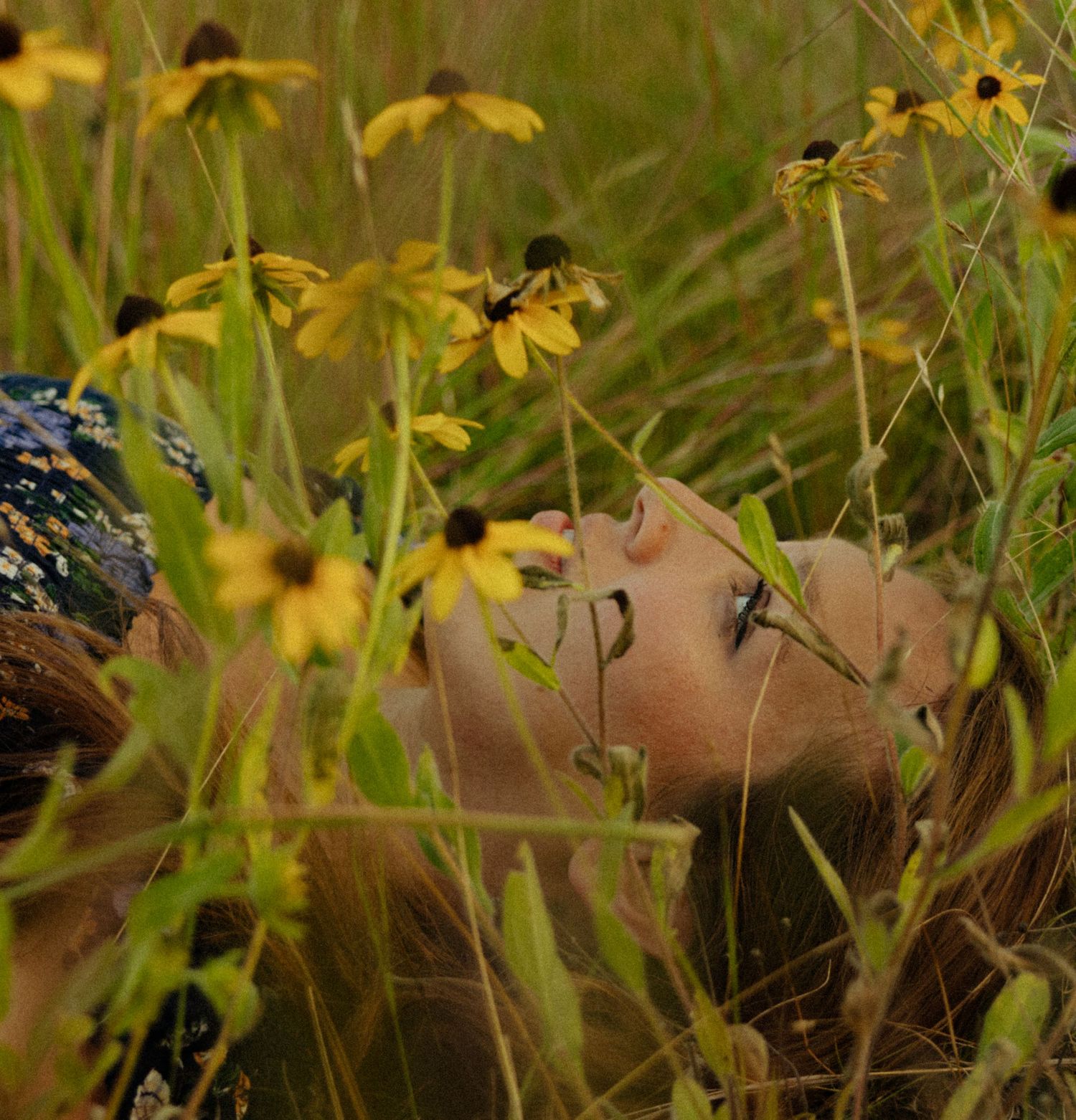 Woman lying among yellow black-eyed Susan wildflowers in a lush green meadow, gazing upward peacefully.