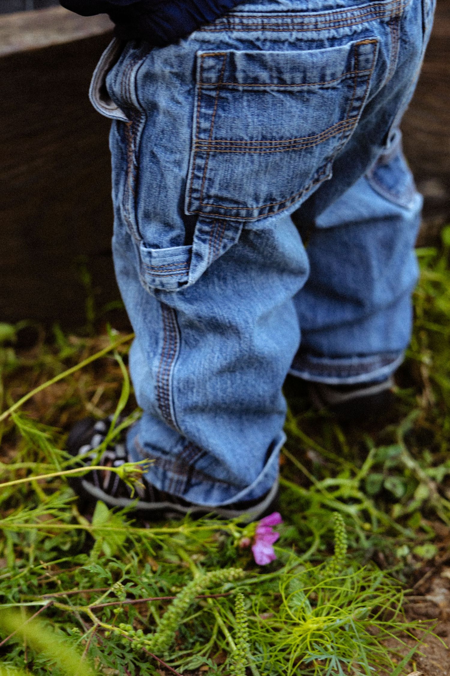 Close-up of a toddler in blue denim overalls and dark shoes standing on green grass with a small pink flower.