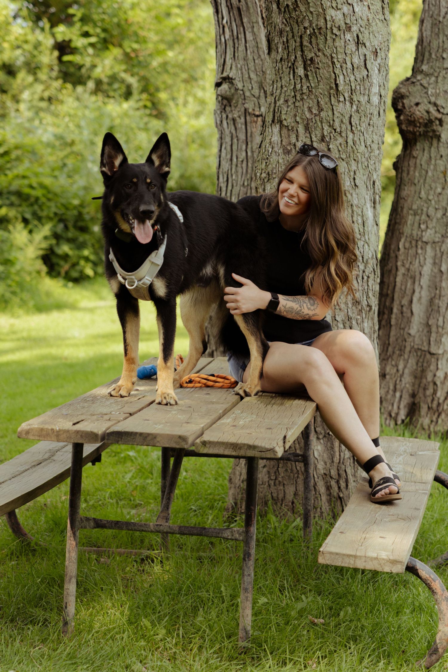 A smiling woman with tattoos sits at a rustic picnic table with a German Shepherd dog in a lush green park.