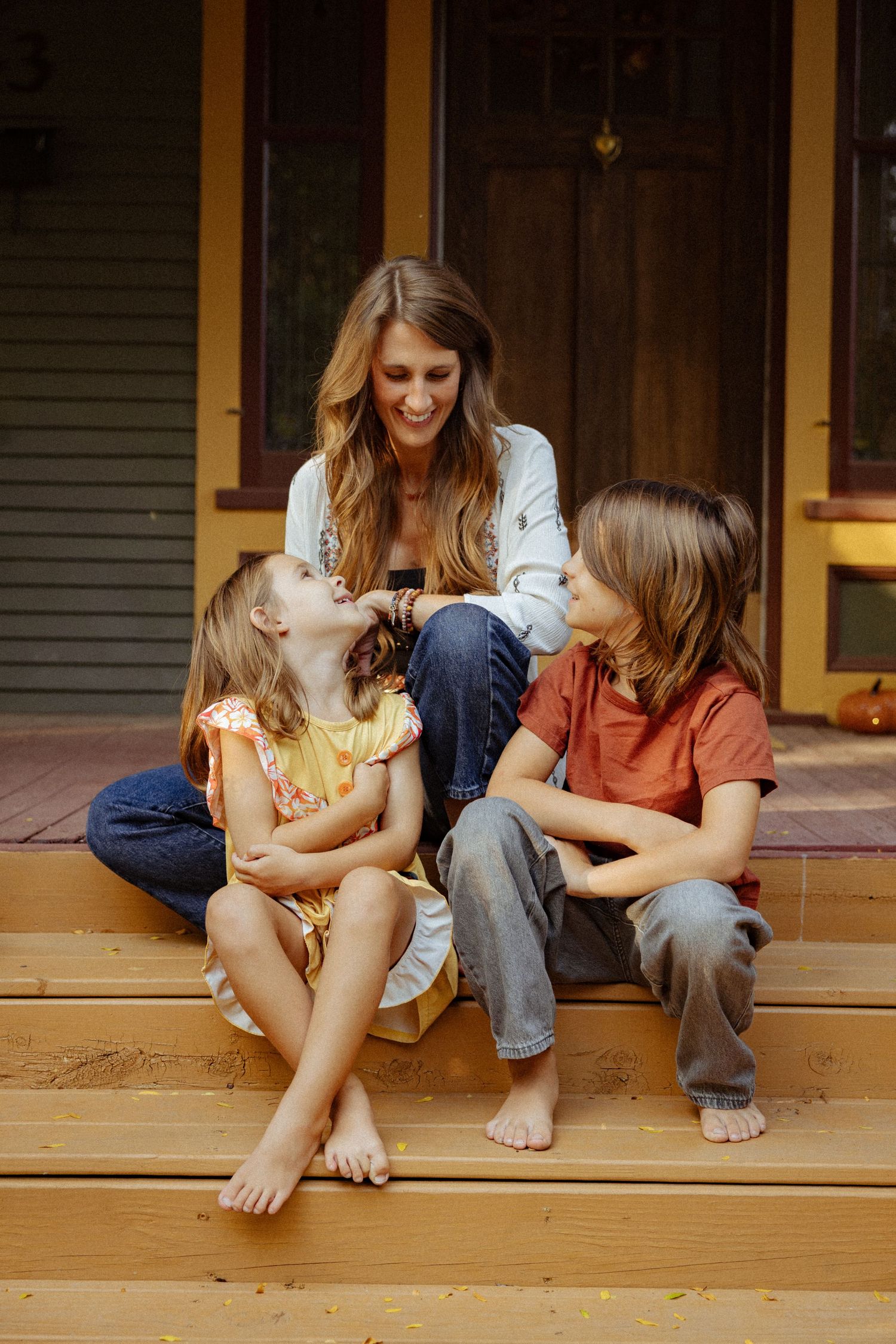 A smiling woman sits on wooden porch steps with two barefoot children, sharing a warm, joyful moment in front of a rustic wooden door.