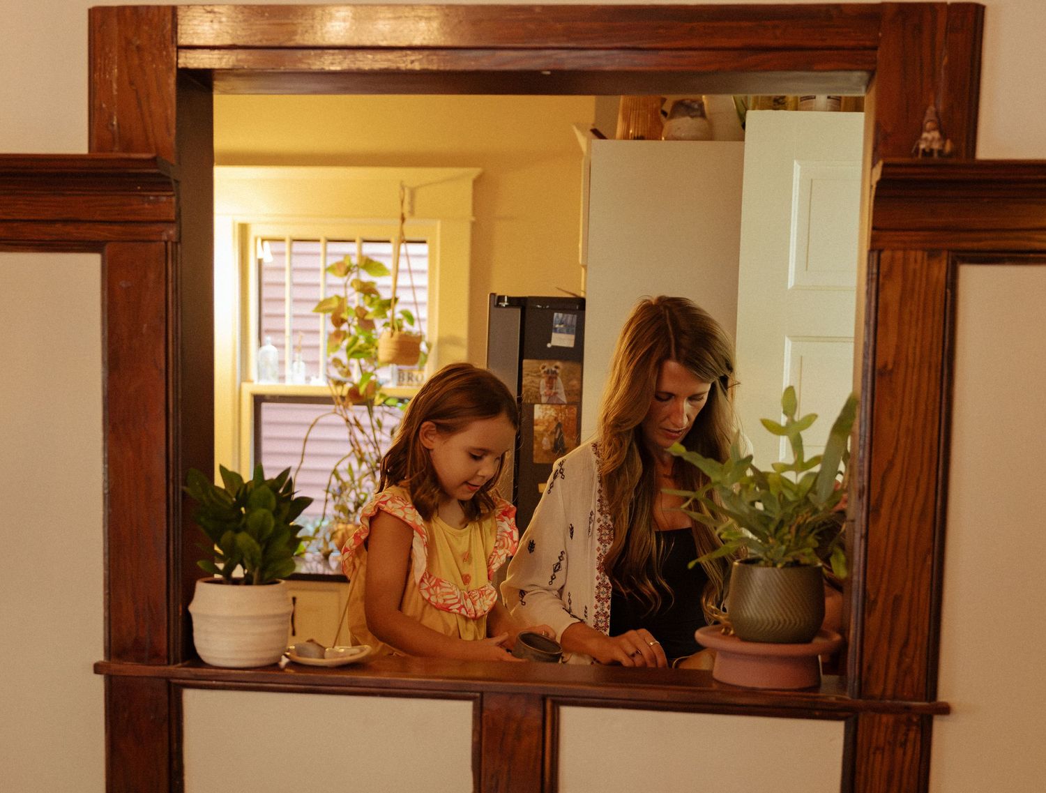 A woman and a young girl stand together in a sunlit kitchen, framed by a wooden pass-through window with potted green plants on the ledge.
