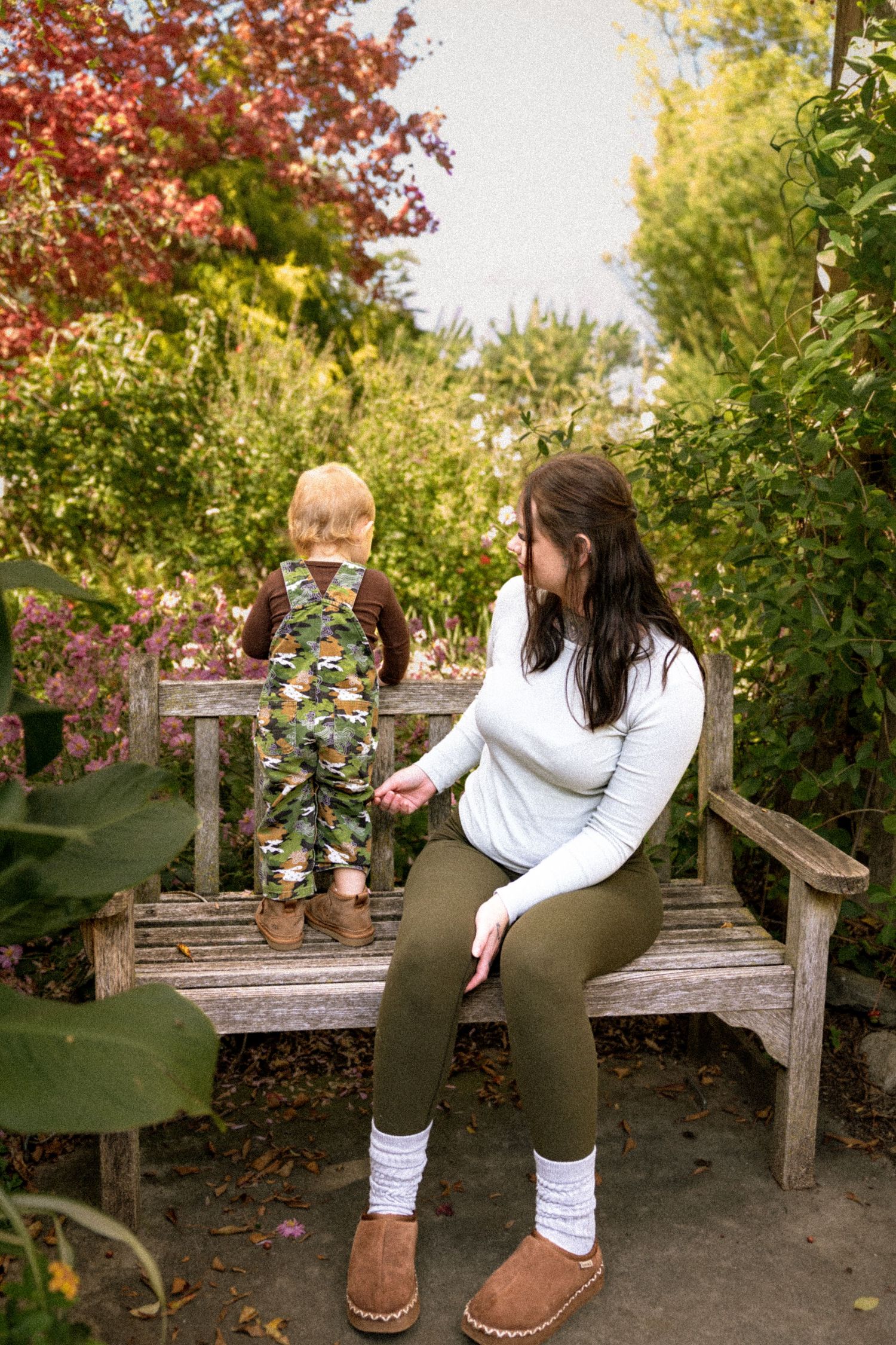 Mother sitting on garden bench watching toddler in camo overalls among colorful autumn foliage.