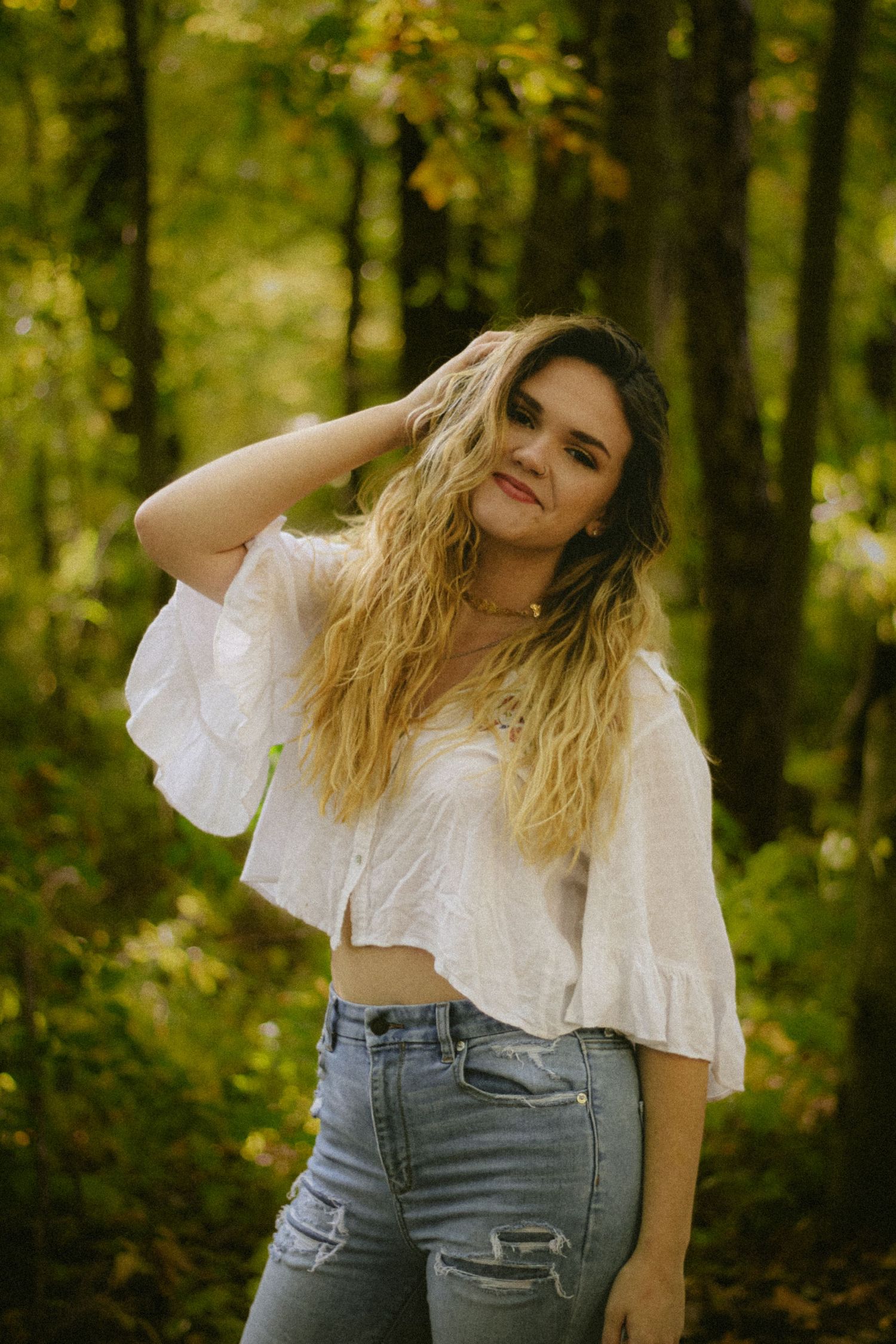 Young woman with wavy blonde hair poses in white crop top and ripped jeans in a lush green forest setting.