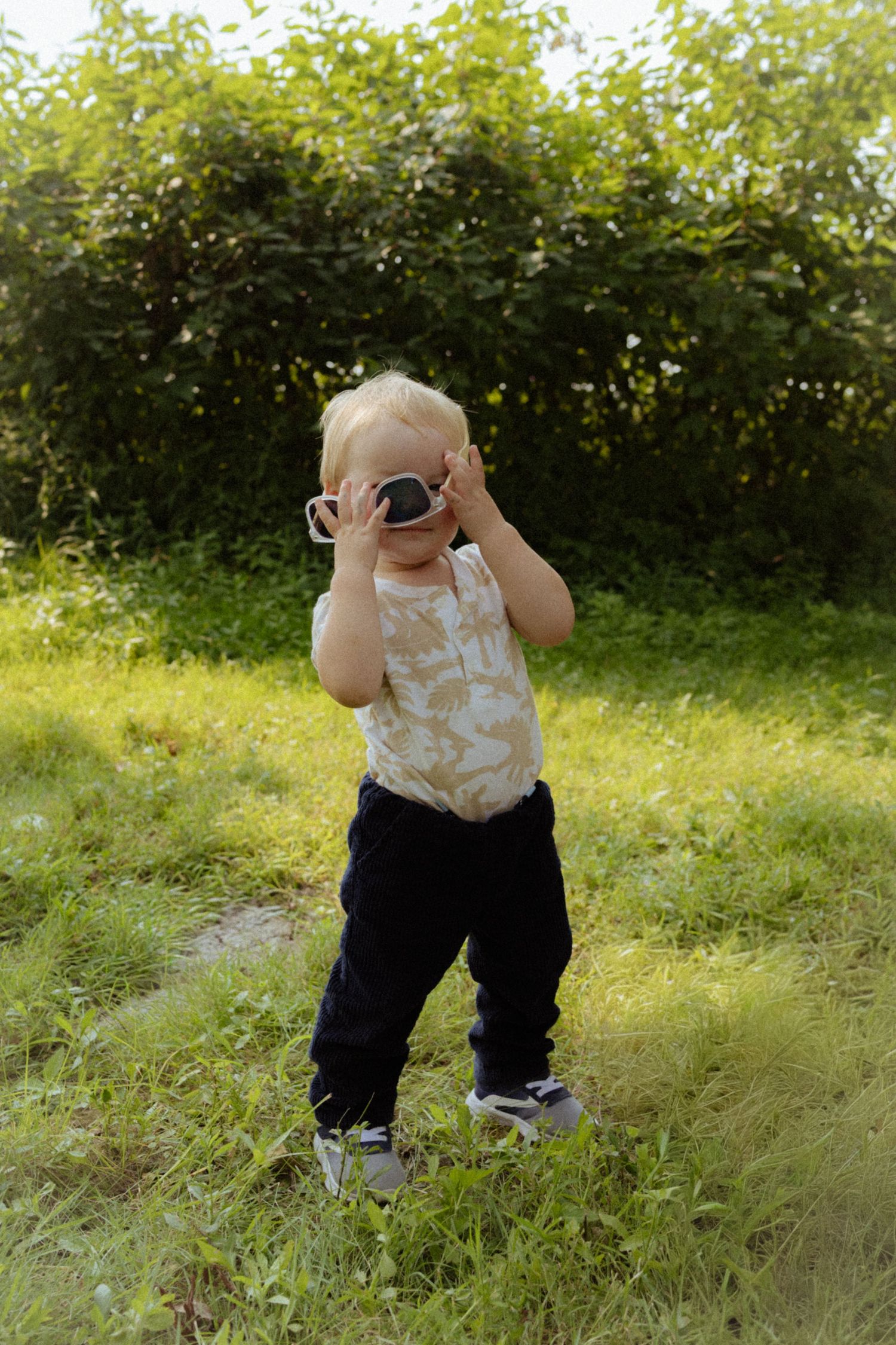 A blonde toddler boy holding sunglasses up to his face while standing on green grass outdoors.