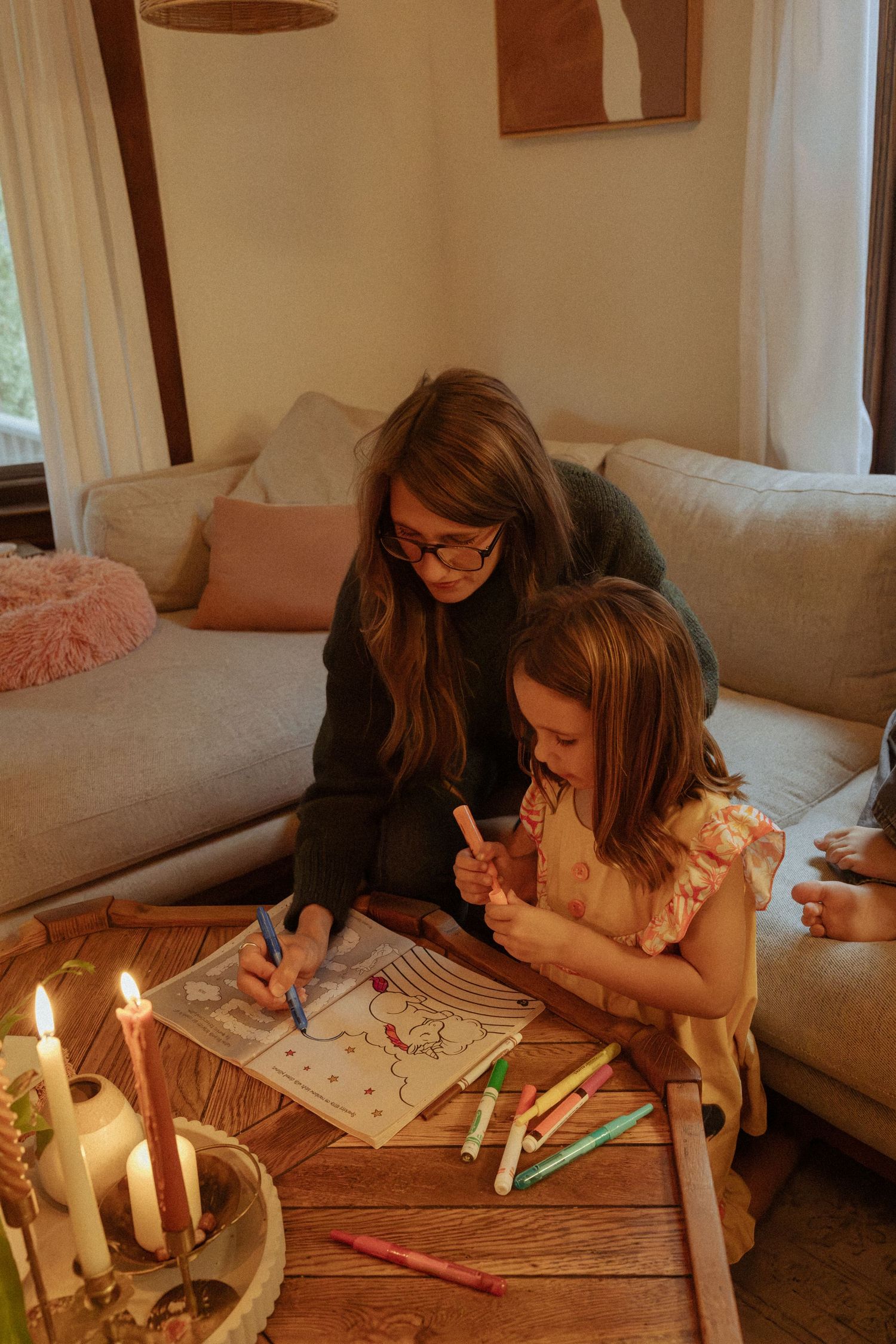 A woman and a young girl color together with markers on a wooden coffee table beside lit candles in a cozy living room.