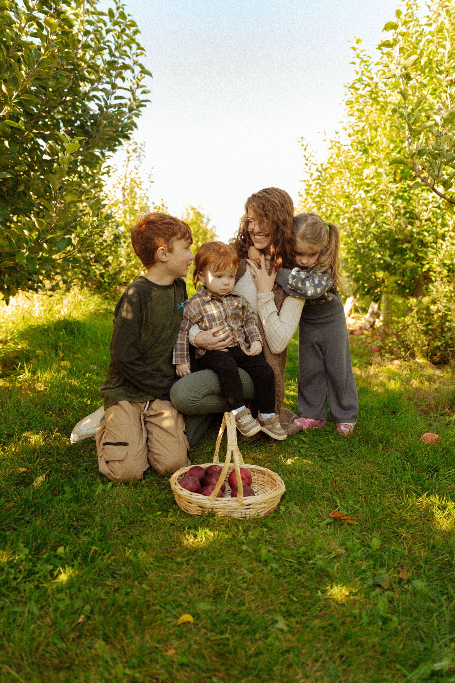 Family picking apples in an orchard, gathered around a wicker basket on green grass.