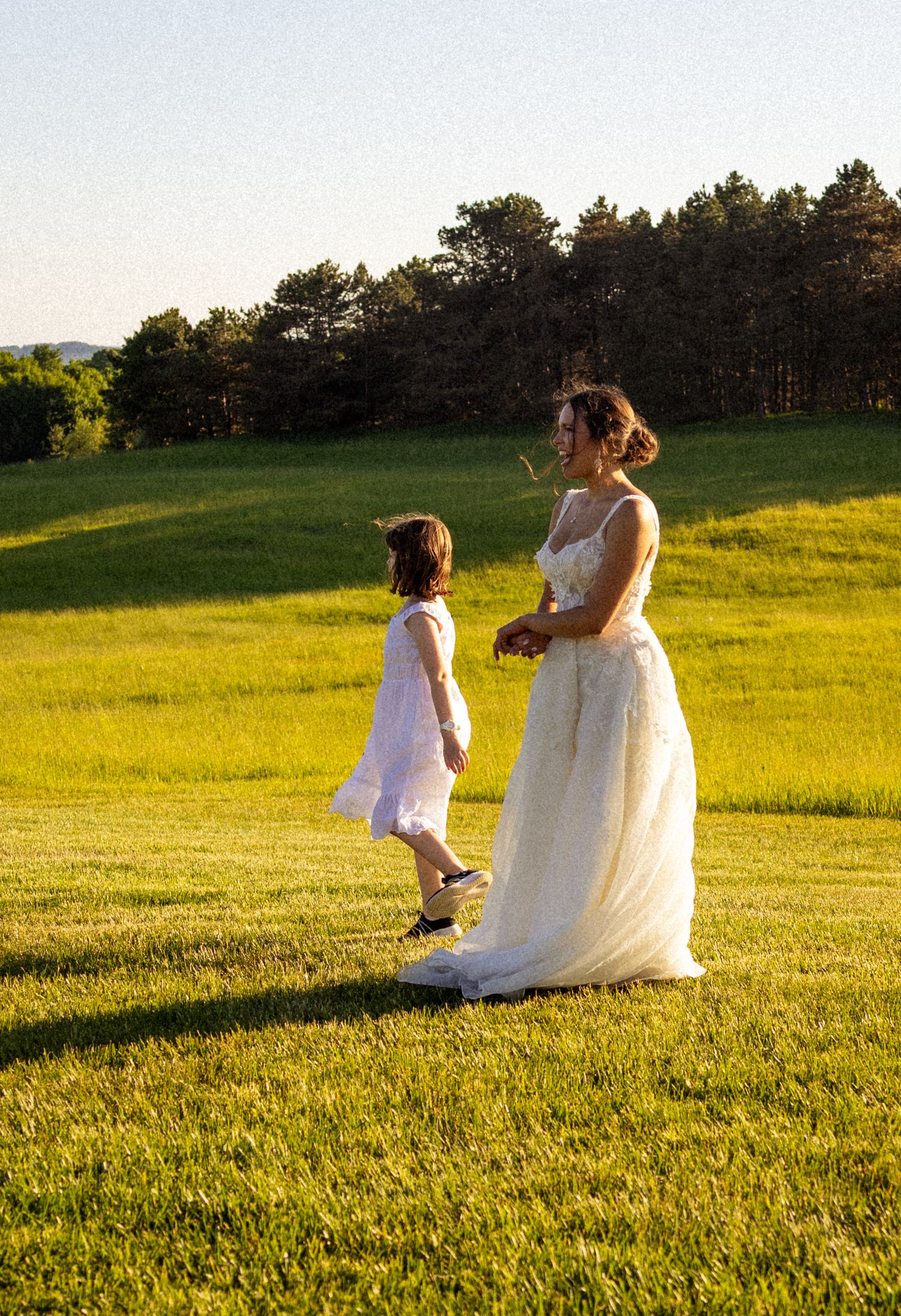 Bride in white gown walks with young flower girl across lush green lawn at golden hour wedding celebration.