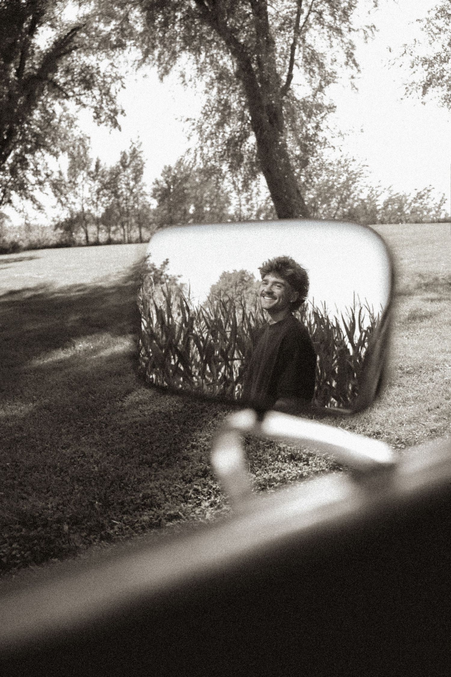 Black and white photo of a smiling person reflected in a car side mirror surrounded by trees and greenery.