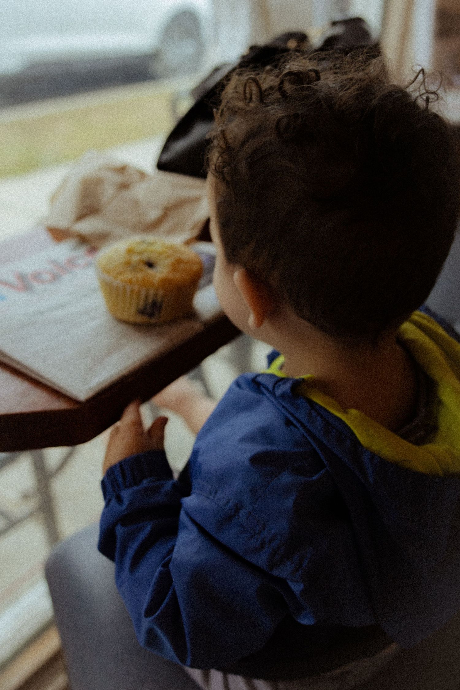 Young child with curly hair sitting at a table looking at a blueberry muffin by a window.