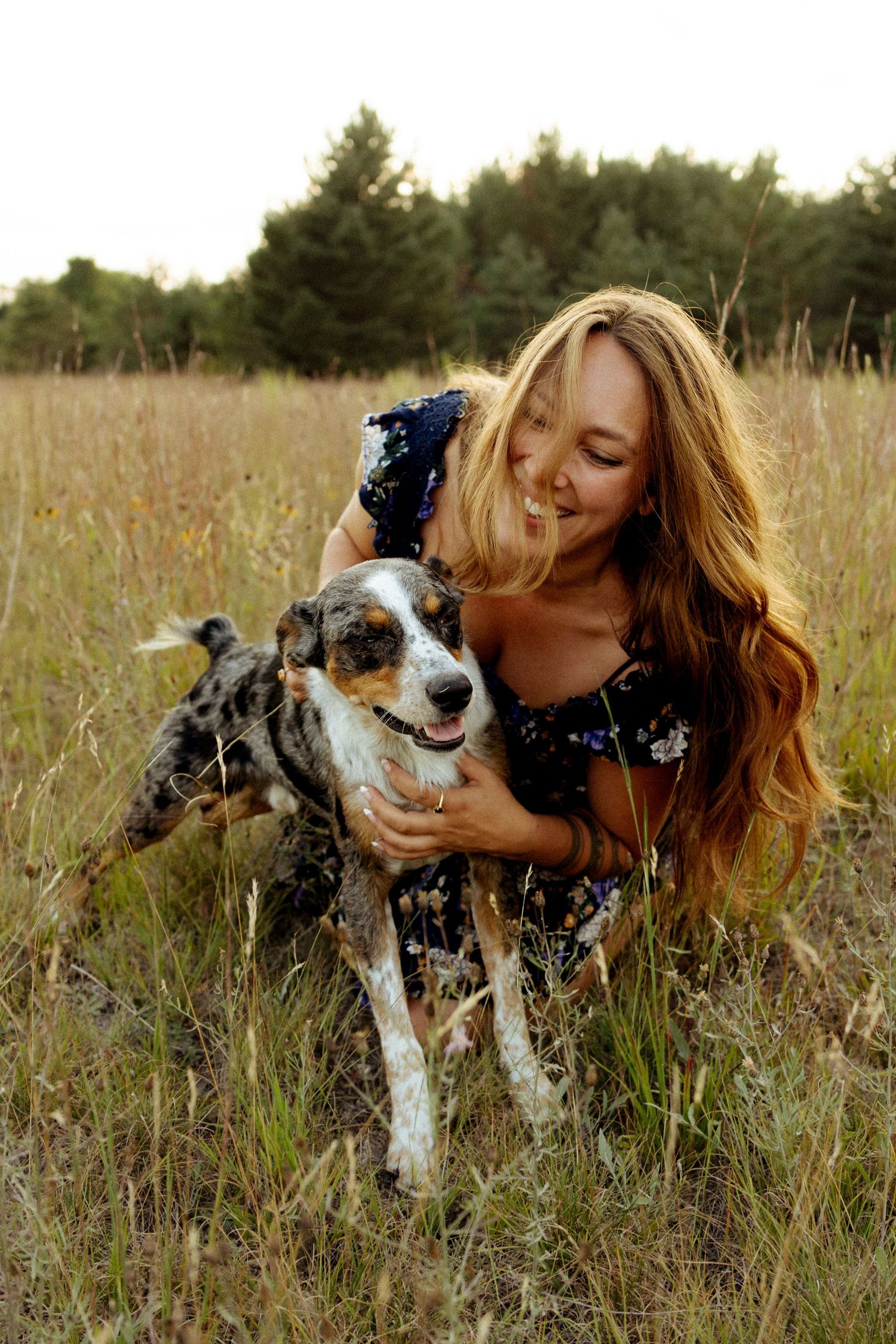 Smiling woman with long blonde hair hugging a spotted dog in a golden field with trees in the background.