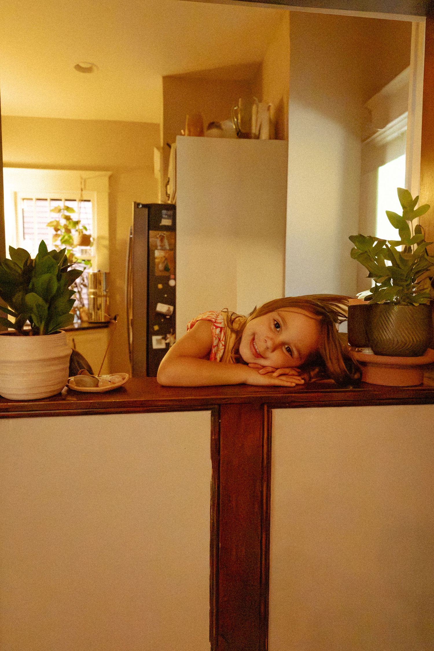 A smiling young girl rests her head on her arms on a wooden kitchen pass-through ledge beside leafy green houseplants.