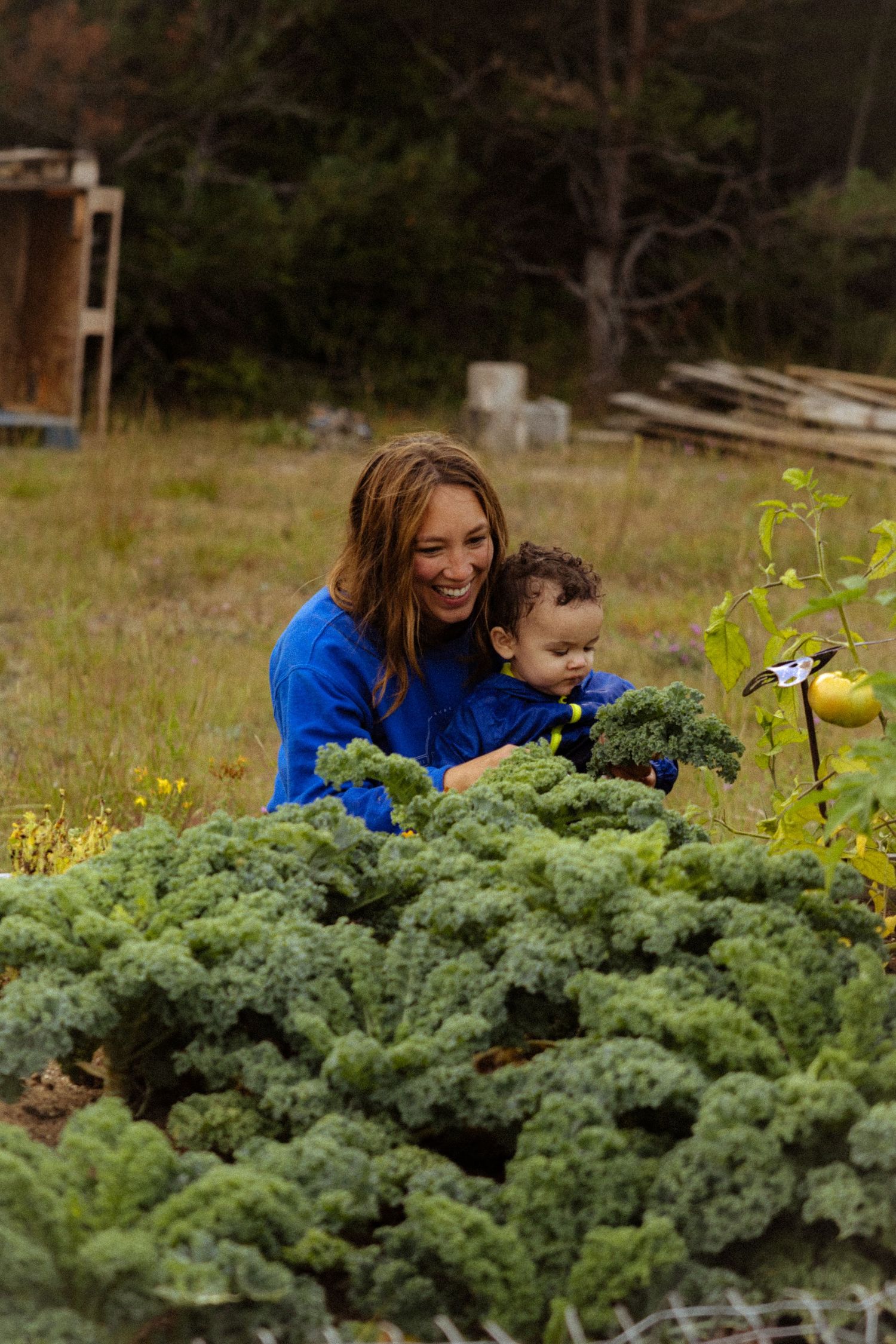 A smiling woman in blue holds a baby near lush kale plants in an outdoor garden setting.