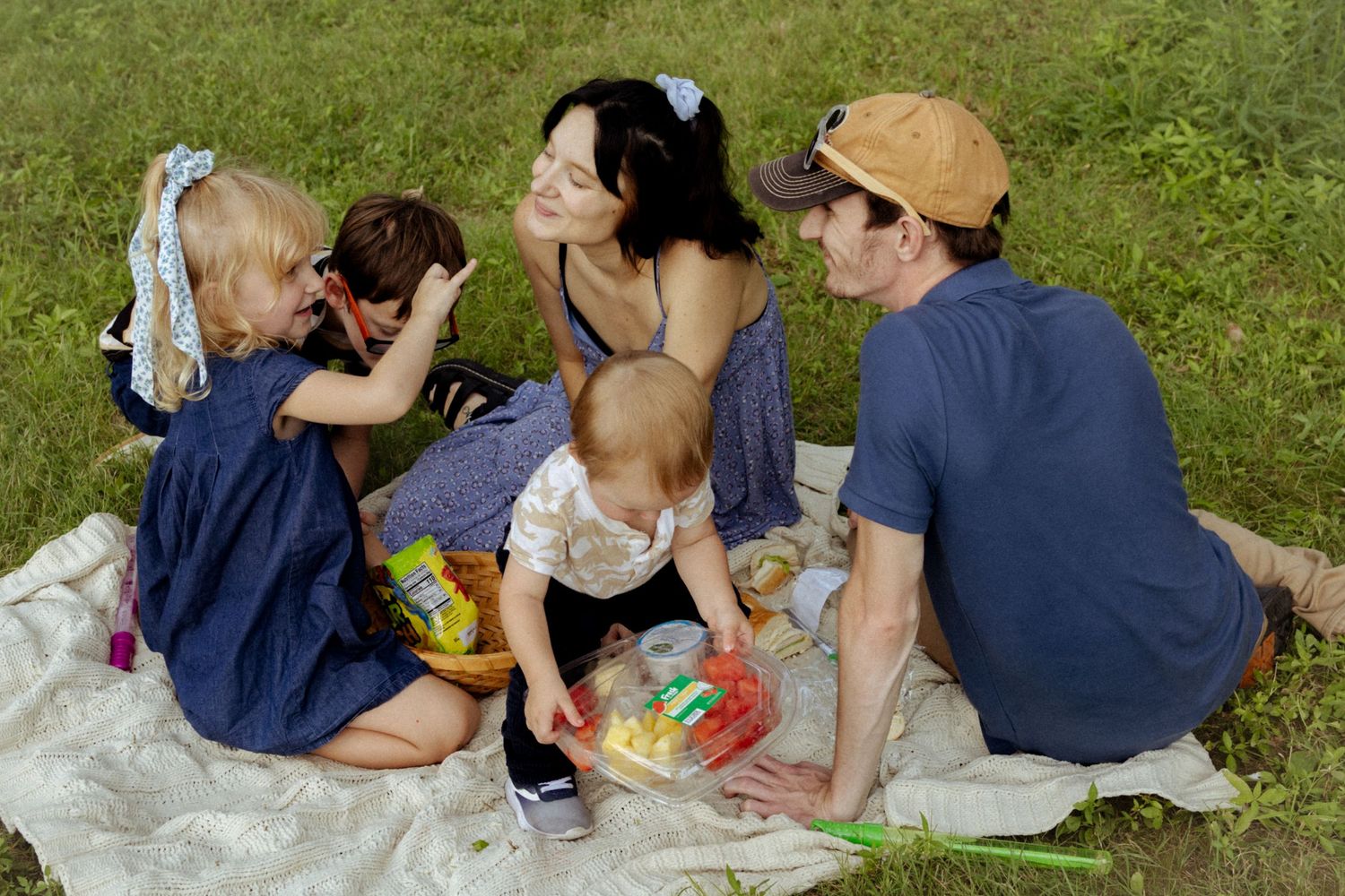 Family of four enjoying a outdoor picnic on a white blanket in a grassy park with snacks and toys.