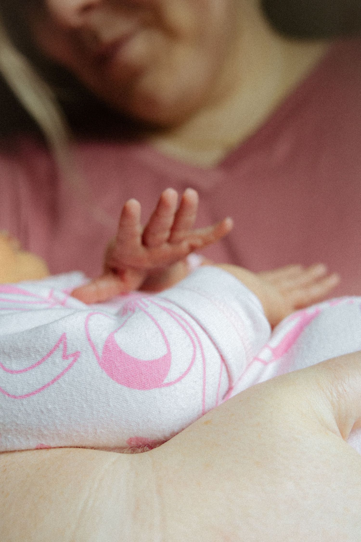 Newborn baby wrapped in white and pink blanket with tiny hands visible, held by mother in pink shirt.