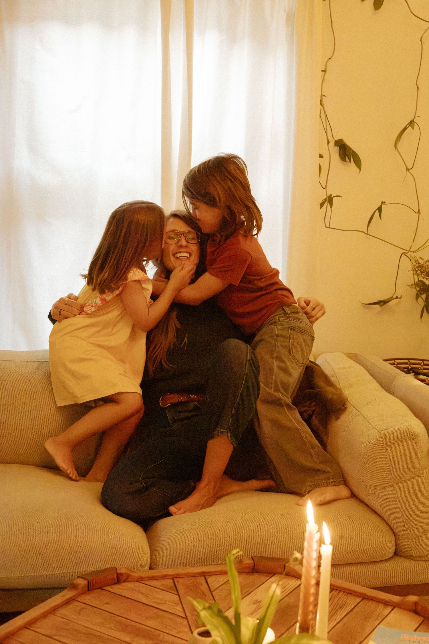 Two children kiss and hug a laughing woman with glasses on a sofa near sheer curtains, with lit candles on a wooden table.