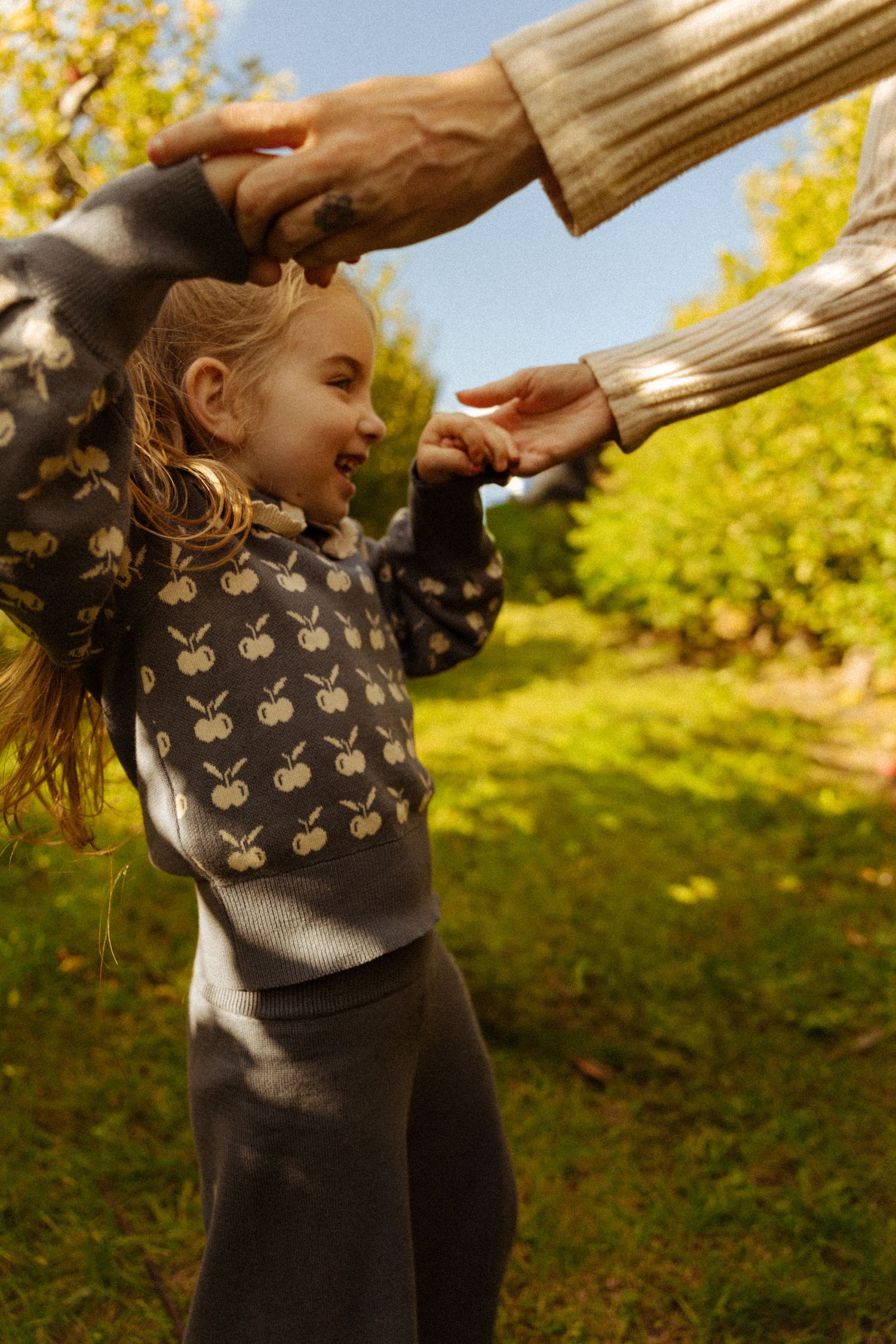 A joyful young girl in an apple-print sweater plays outdoors, holding hands with an adult in a sunny green garden.