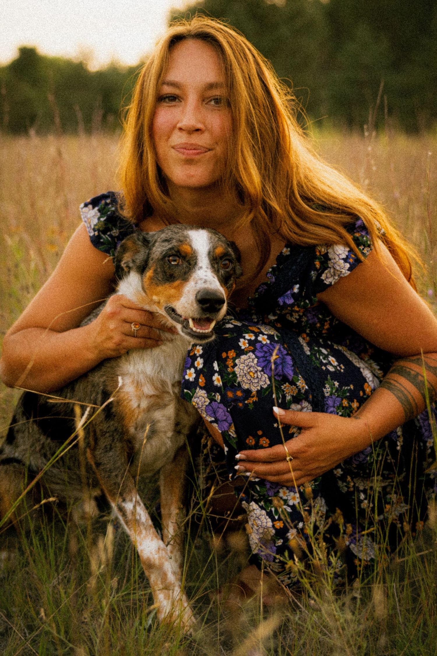 Woman with long auburn hair in floral dress hugging a merle Australian Shepherd dog in a golden grass field.