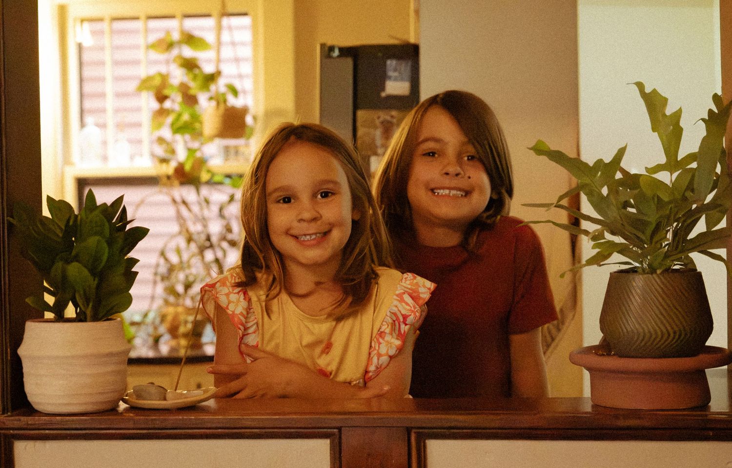 Two smiling young girls lean on a wooden counter in a warm kitchen, surrounded by green potted plants.