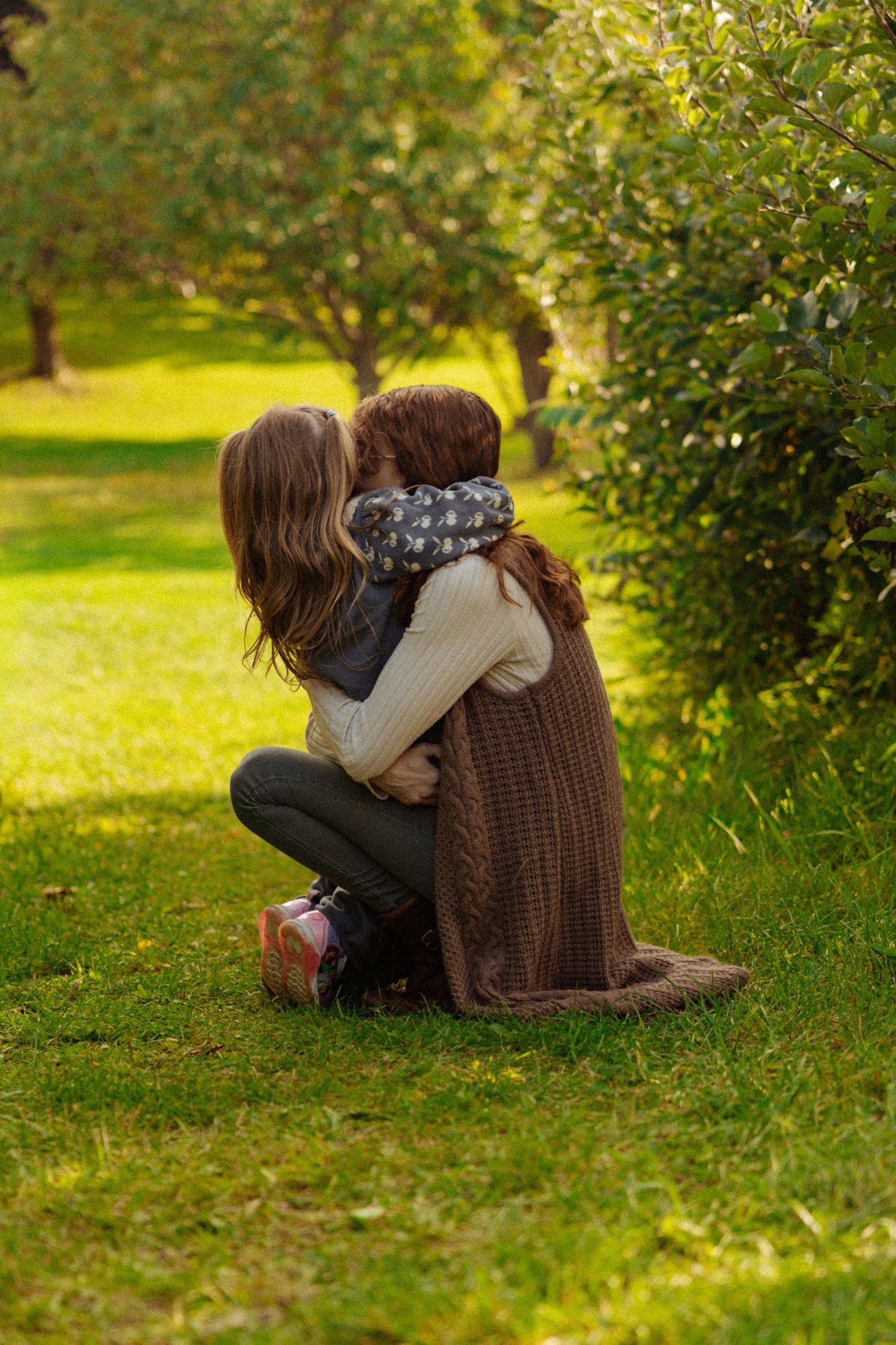 A mother and child share a tender embrace while sitting on green grass in a sunlit garden.