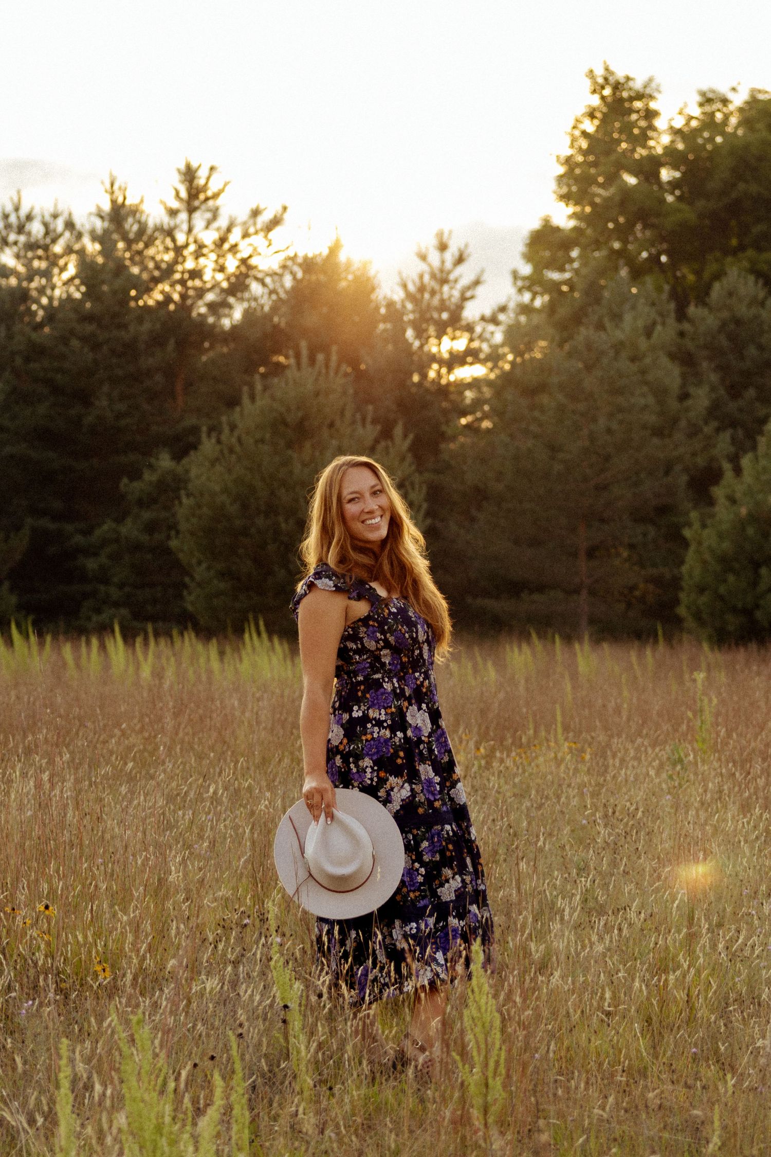 Smiling woman in floral dress holding a hat in a golden field at sunset with trees in the background.