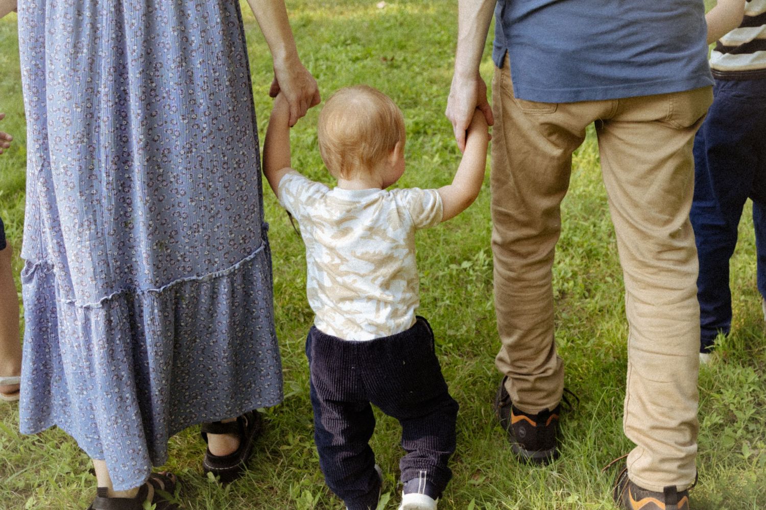 A toddler holds hands with parents while taking first steps on green grass outdoors.