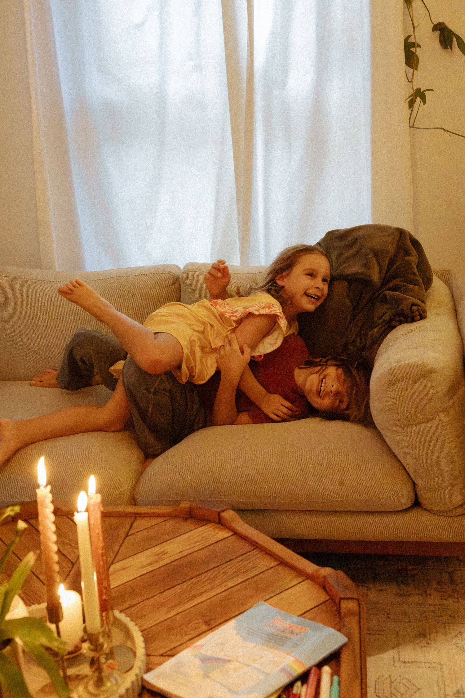 Two laughing children tumble playfully on a beige sofa behind a wooden coffee table with lit candles and coloring supplies.